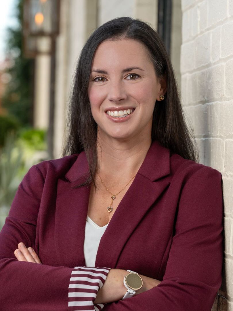 A woman in a maroon jacket is leaning against a brick wall with her arms crossed.