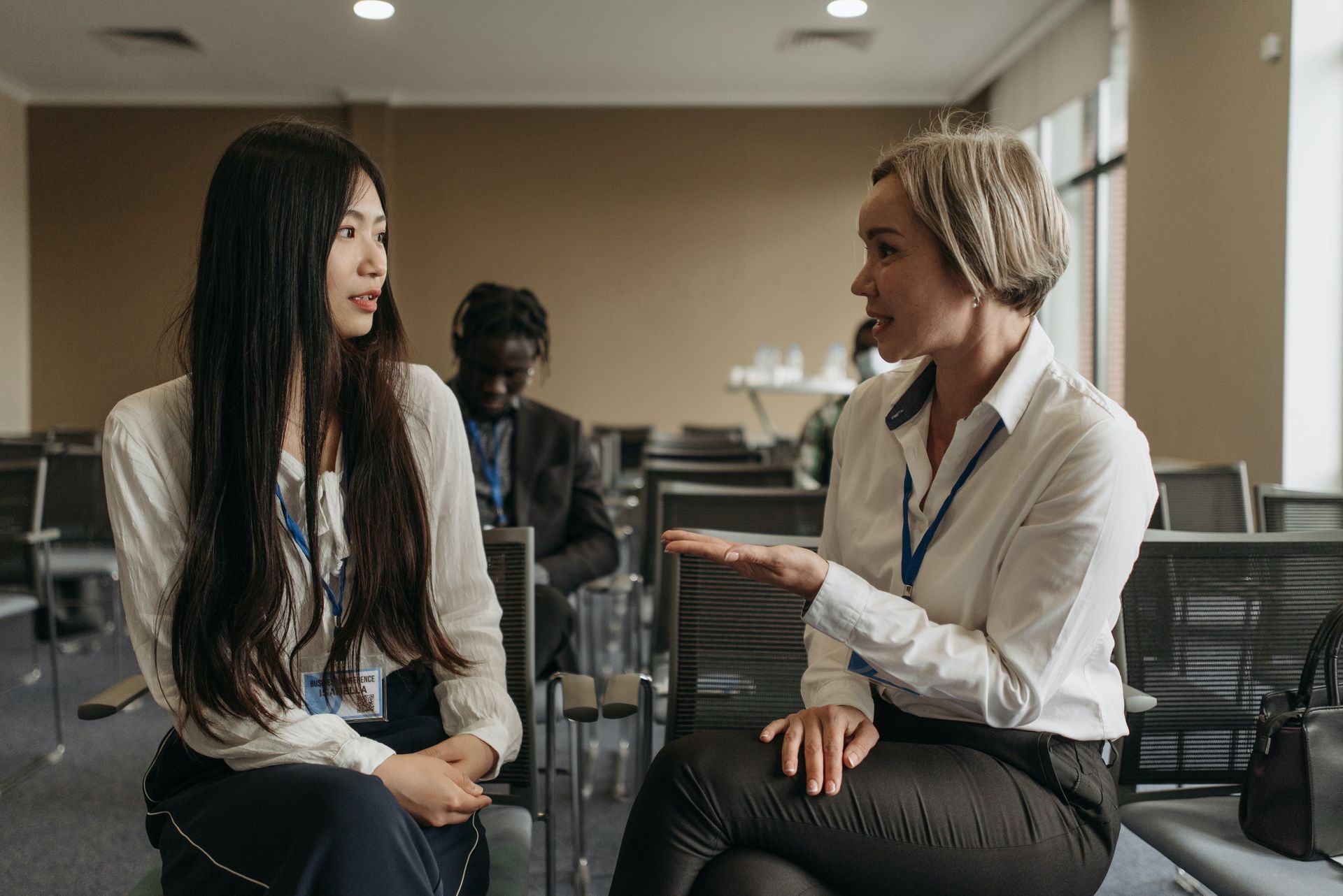 Two women are sitting next to each other in a room talking to each other.