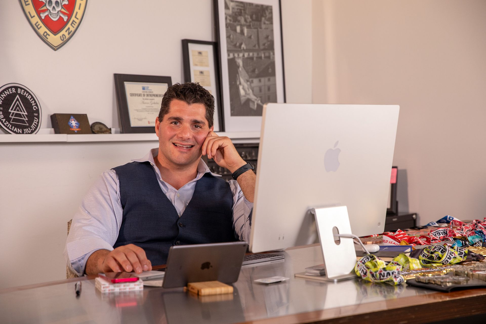 Man sitting at a desk with computer, smiling, wearing a vest. Office setting, accessories visible.