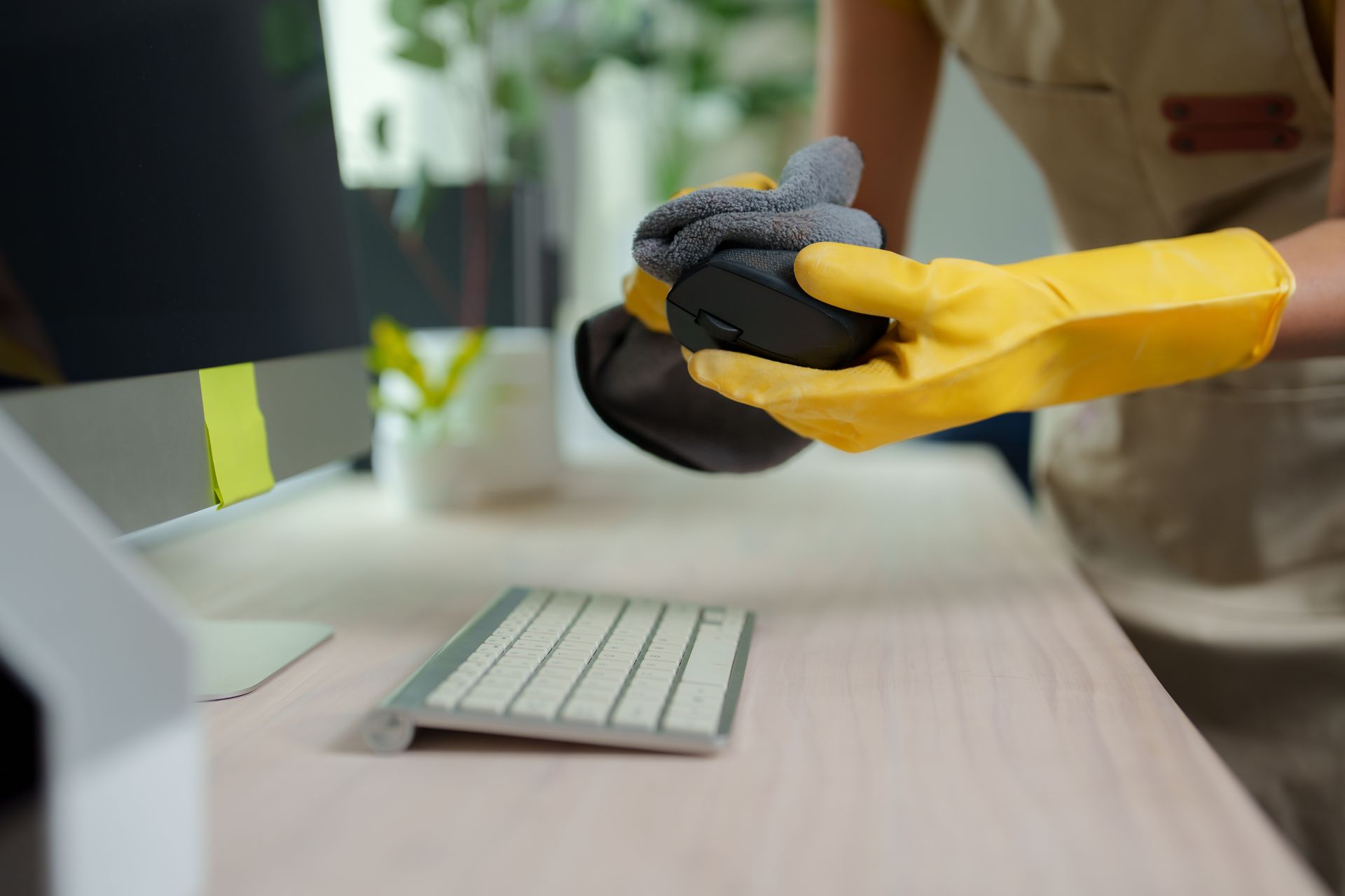 A person wearing yellow rubber gloves wipes a computer mouse with a grey cleaning cloth at a tidy desk.