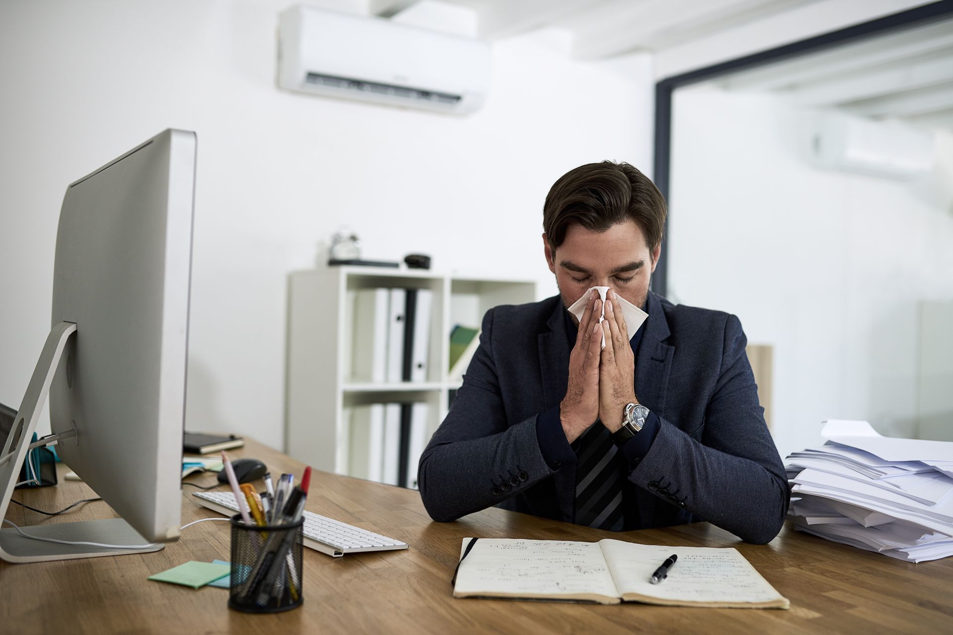 Man in suit, blowing nose at desk in office.