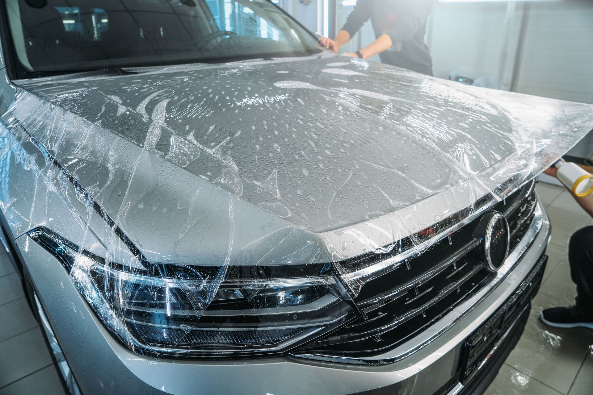 Two people applying clear protective film to a silver car's hood in a service bay.