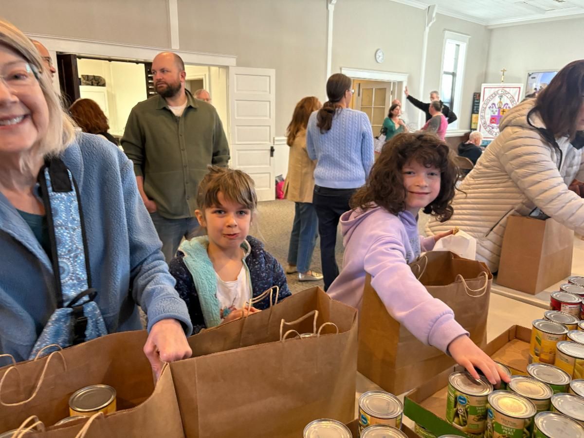 People packing food into bags at a food bank. Two children smile, holding the bags. Canned goods are on a table.