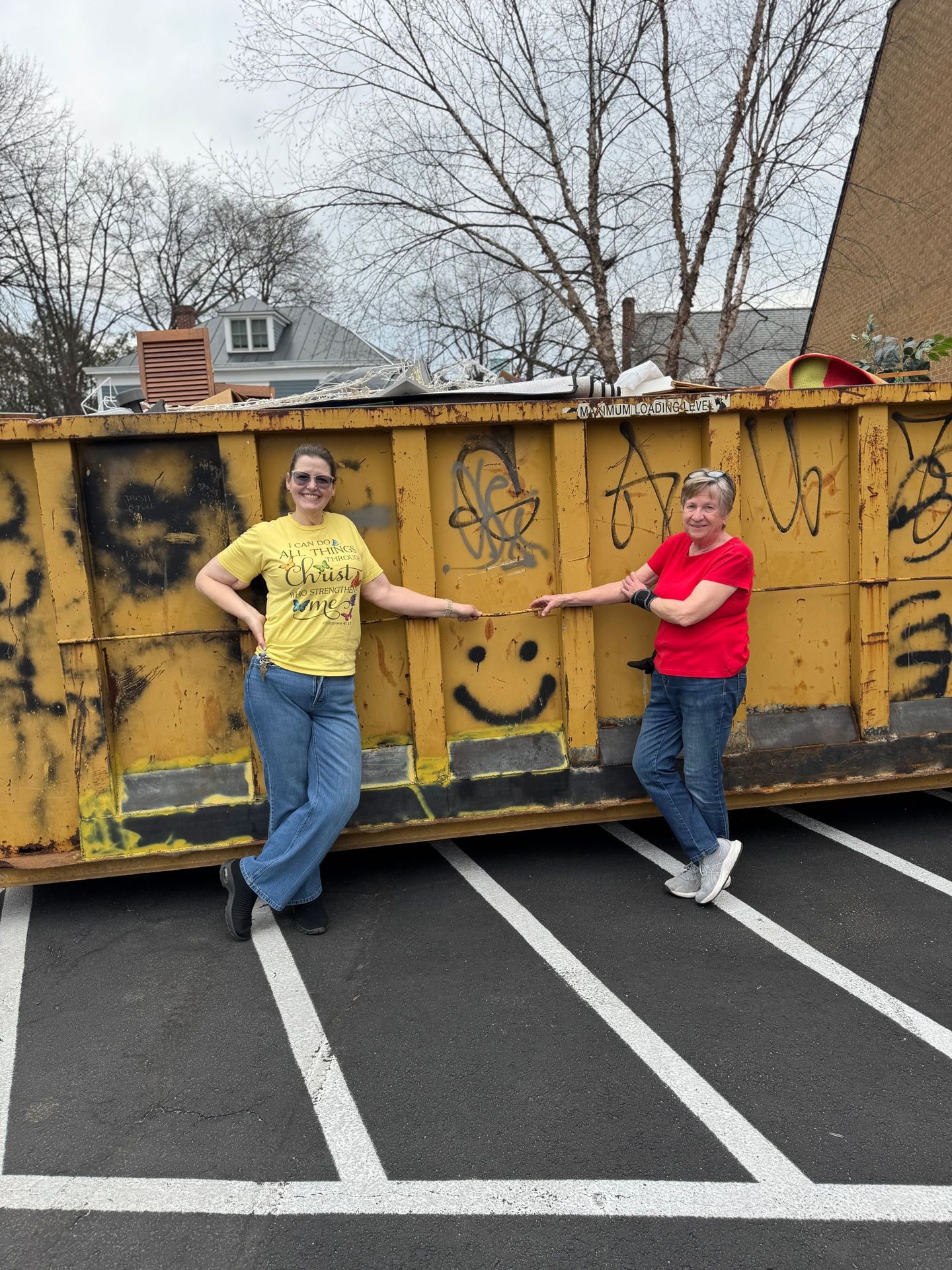 Two people pose beside a yellow dumpster with graffiti in a parking lot. One wears yellow, the other red.