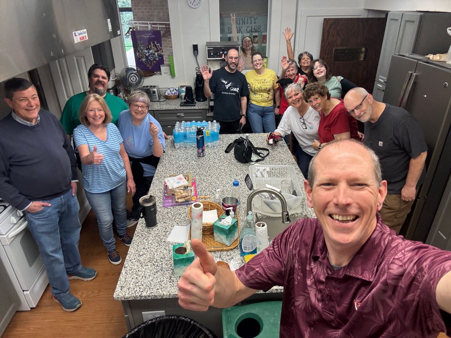 Group of people in a kitchen smiling and giving thumbs up. A man takes a selfie.