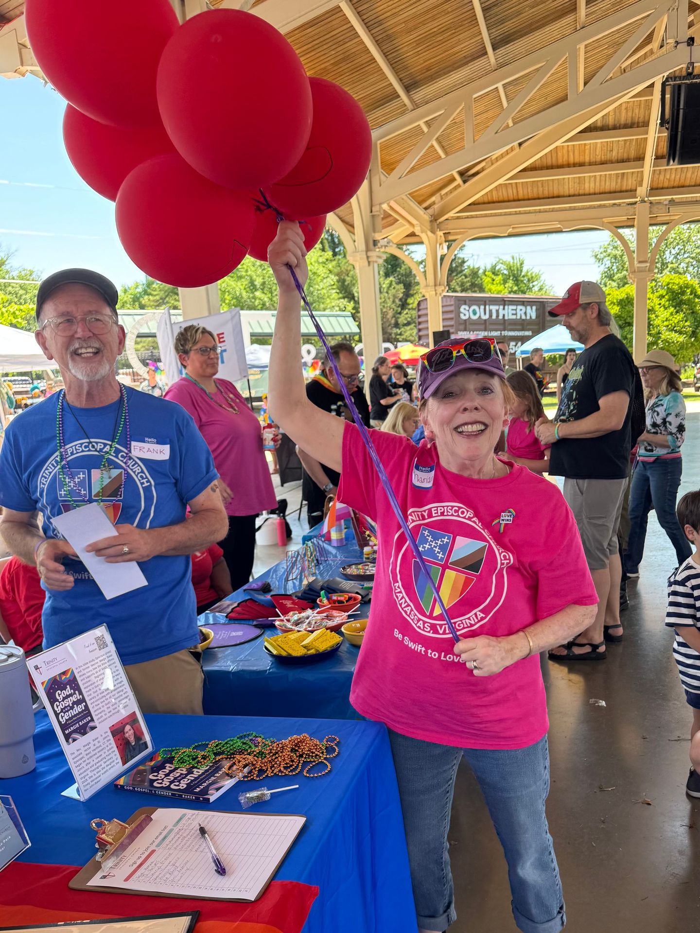 Woman holding red balloons at a booth, smiling. Others in the background. Blue and red color scheme.