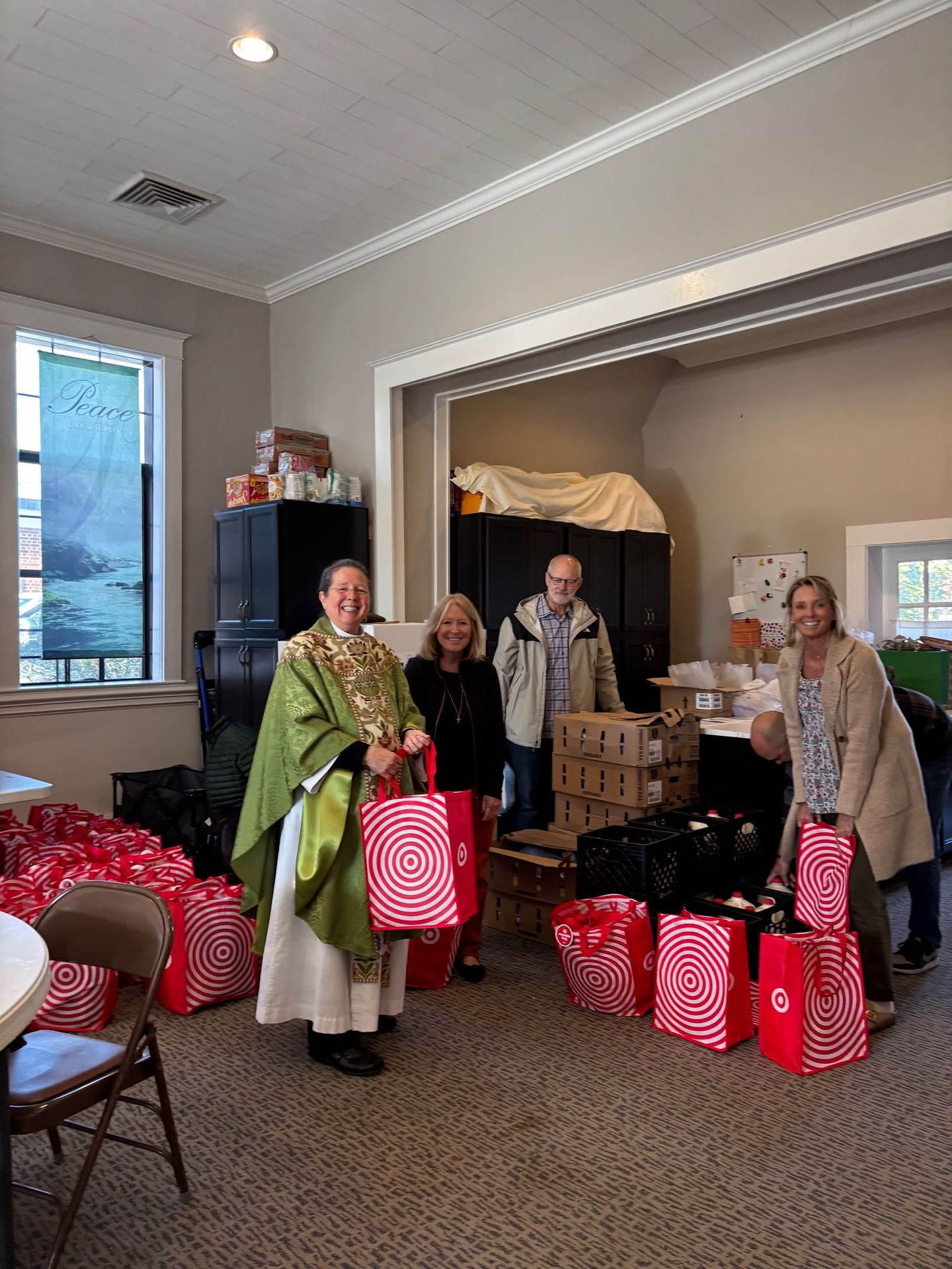 Group of people holding gift bags, smiling in a room. Priest in green vestment stands among red wrapped gifts.