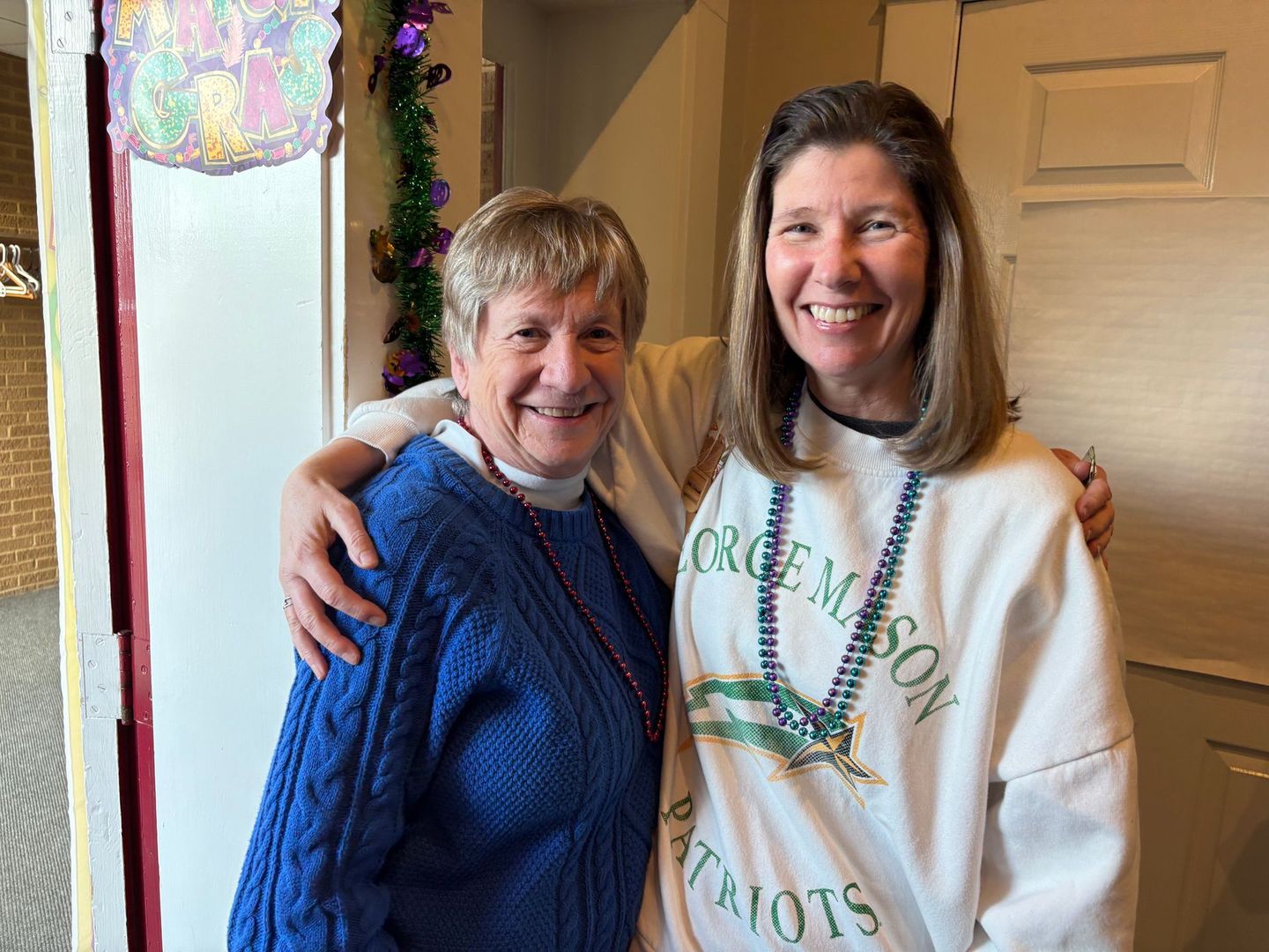 Two women with Mardi Gras beads, smiling, indoors. One in blue sweater, the other in a George Mason sweatshirt.