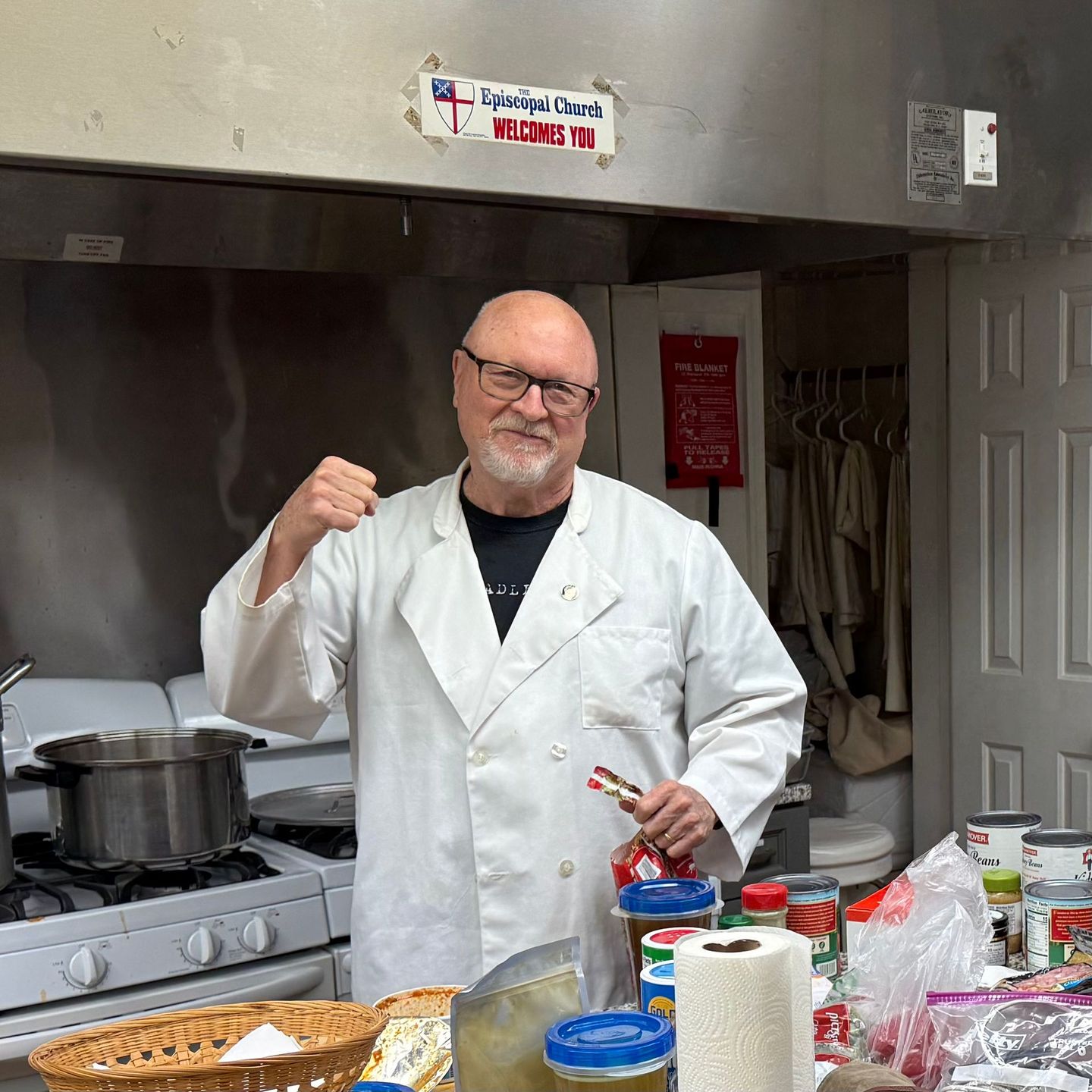 Man in chef coat, kitchen. Holds bottle, fist raised. Pots on stove, food prep area.