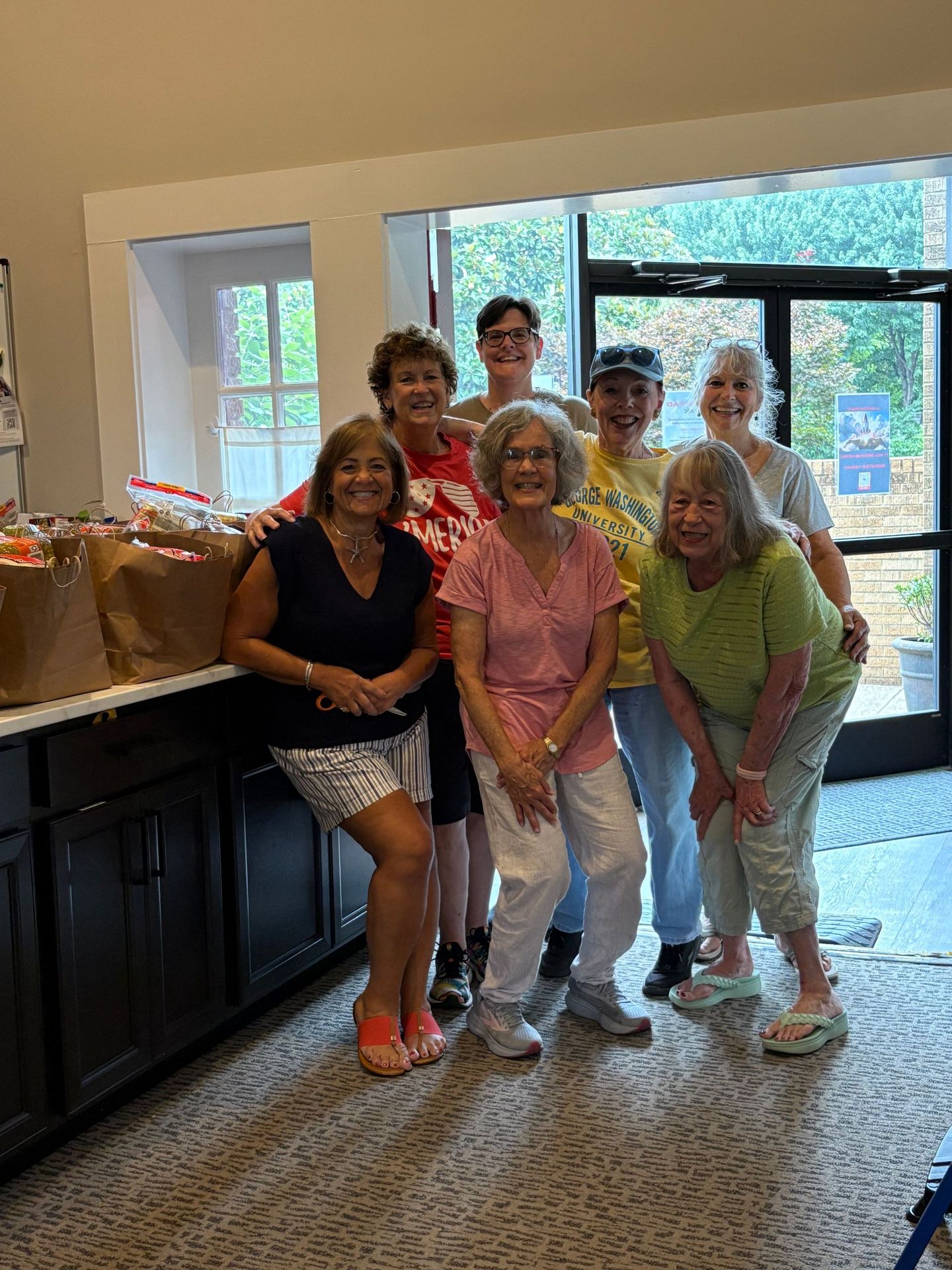 Group of seven people smiling, standing near a counter with brown paper bags. Interior setting with a door and window.