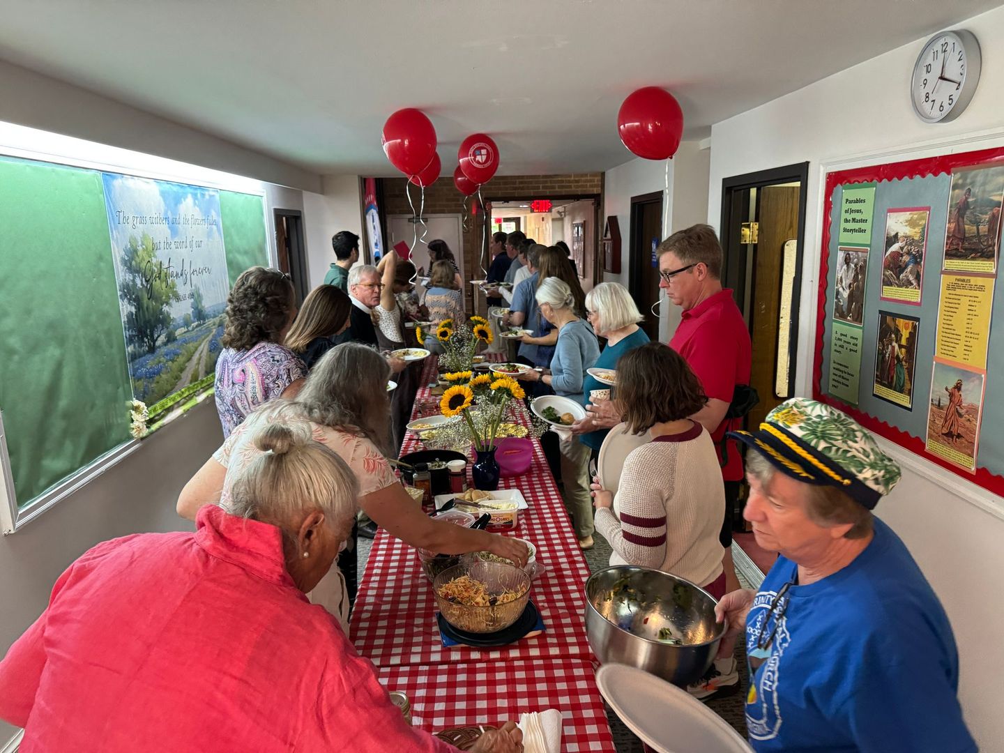 People gather around a long table with food. Red balloons and sunflowers decorate the space.