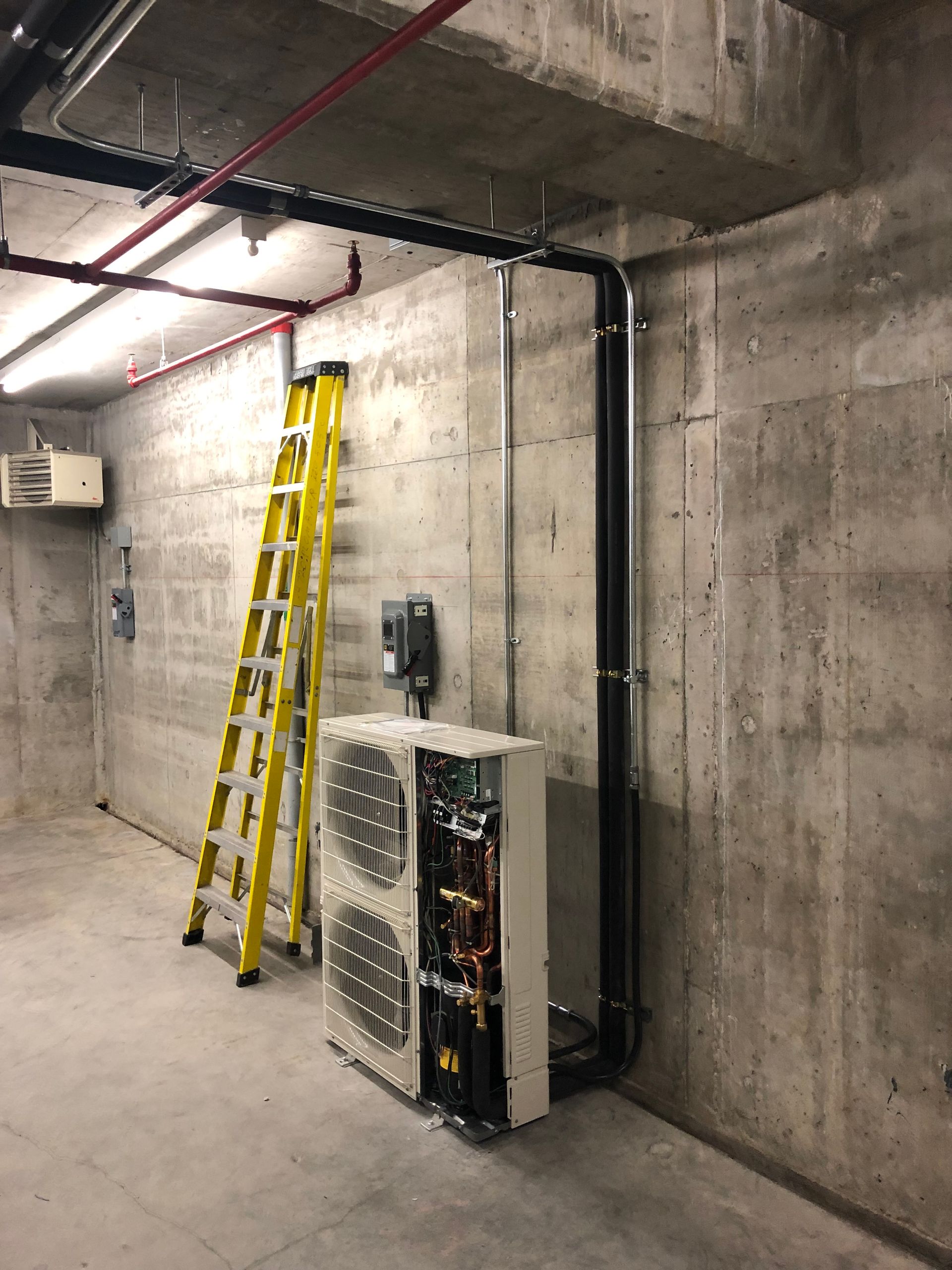 Une échelle jaune est appuyée contre un mur en béton à côté d'une unité de pompe à chaleur avec des tuyaux exposés.