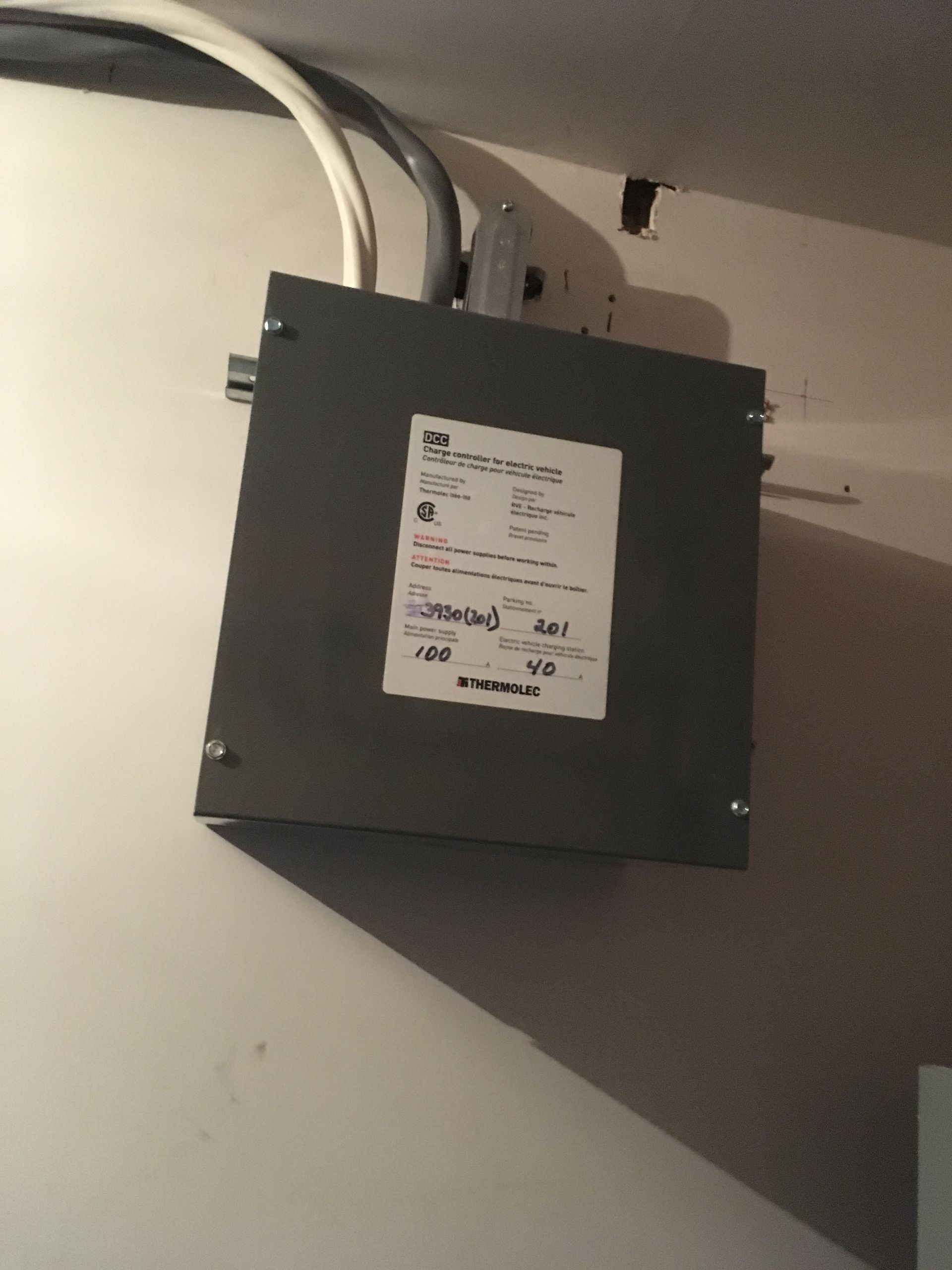 Un transformateur électrique gris monté sur un mur blanc avec un conduit et une étiquette.