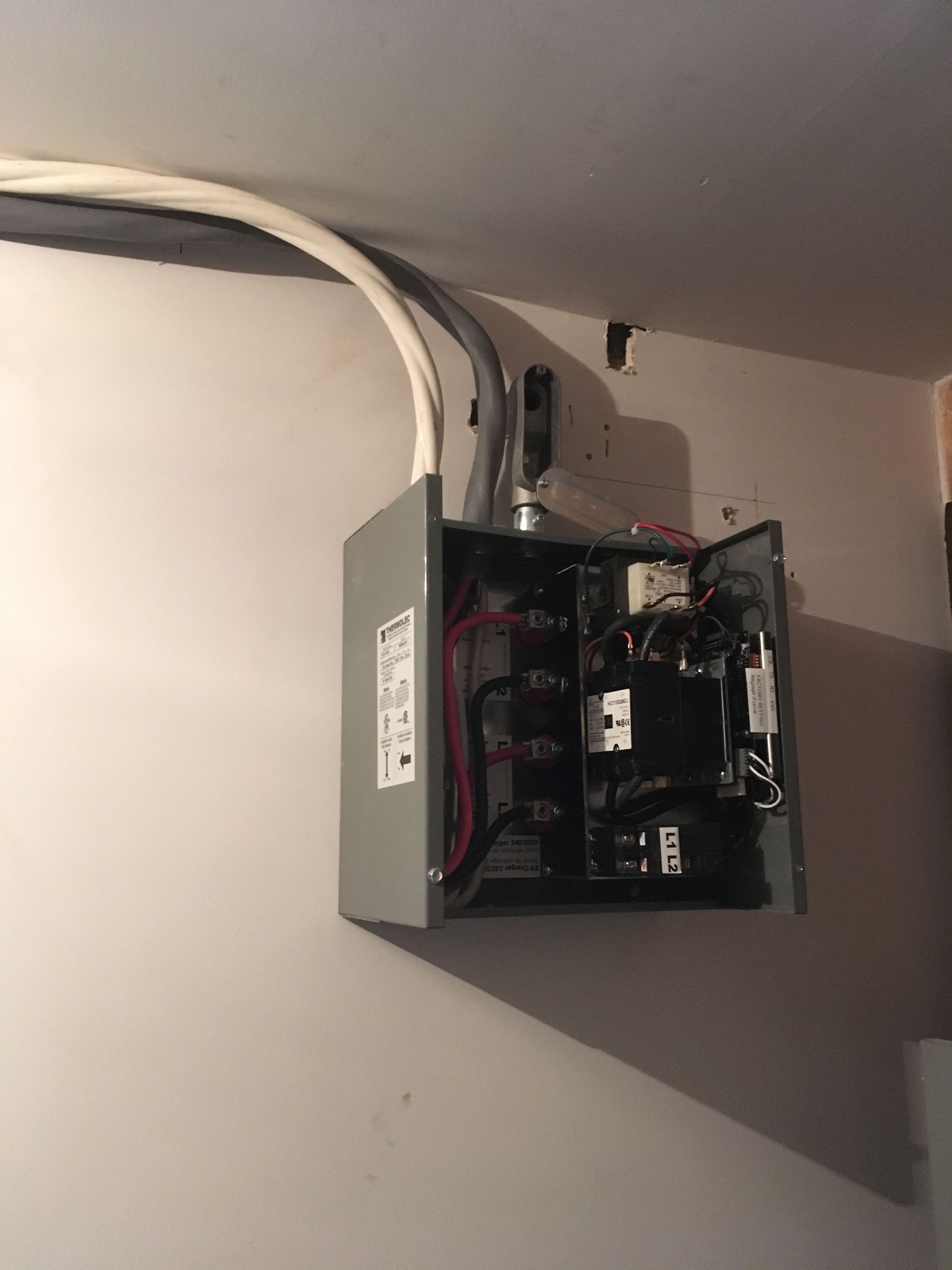 Panneau électrique gris monté sur un mur blanc, porte ouverte, montrant les disjoncteurs et le câblage. Conduit au-dessus.