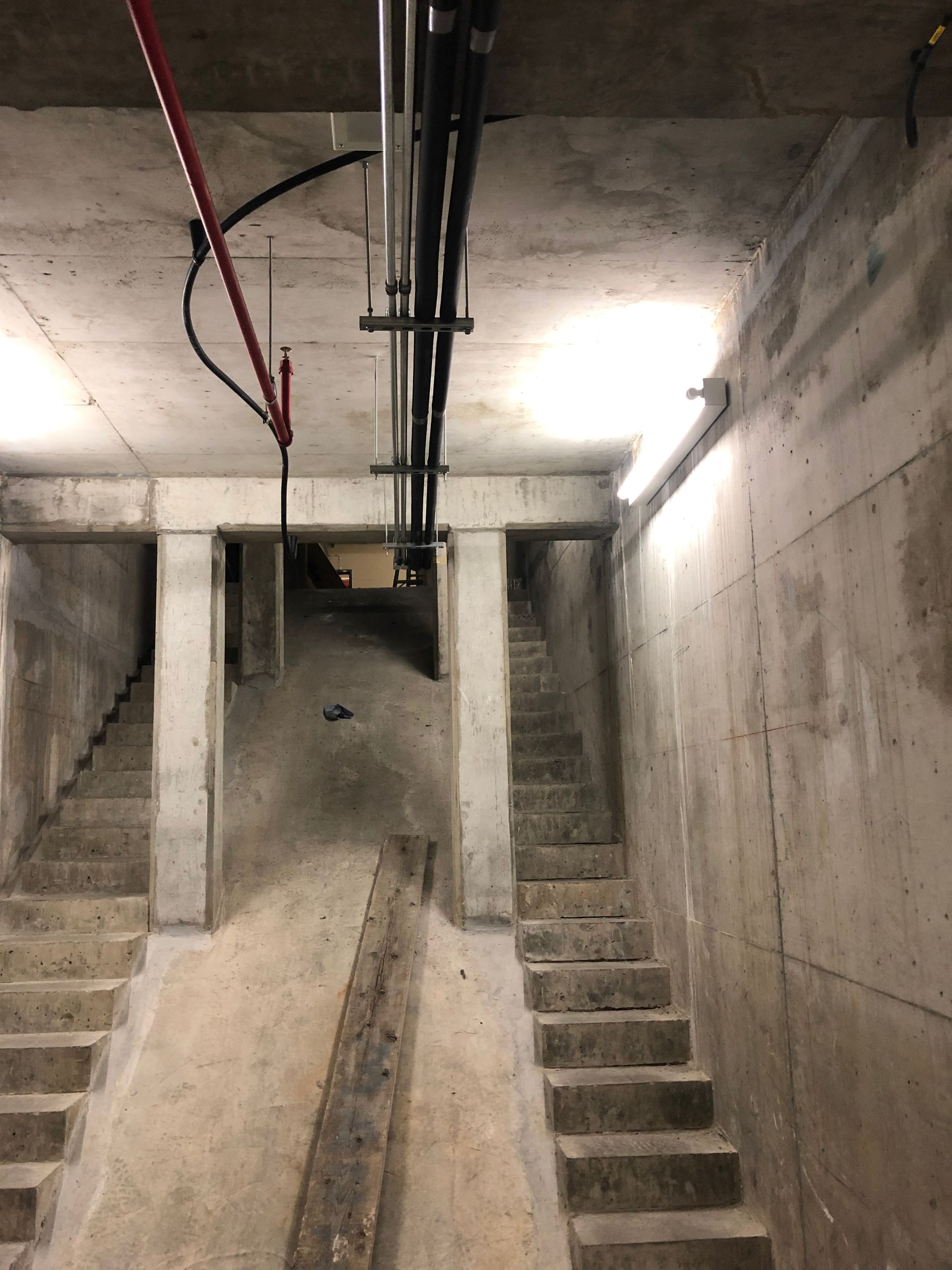 Escalier en béton menant vers un espace brut et inachevé avec des tuyaux et un éclairage apparents.