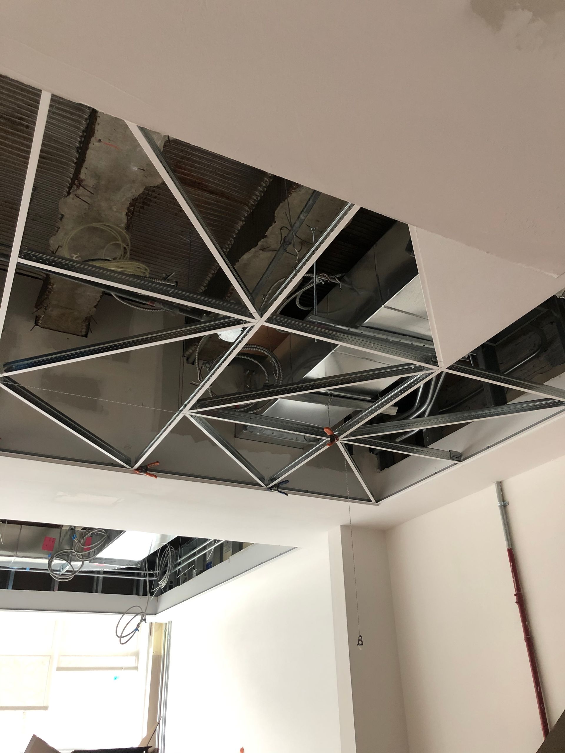 Plafond en construction avec grille métallique triangulaire exposant tuyaux et câblage. Murs et moulures blancs.