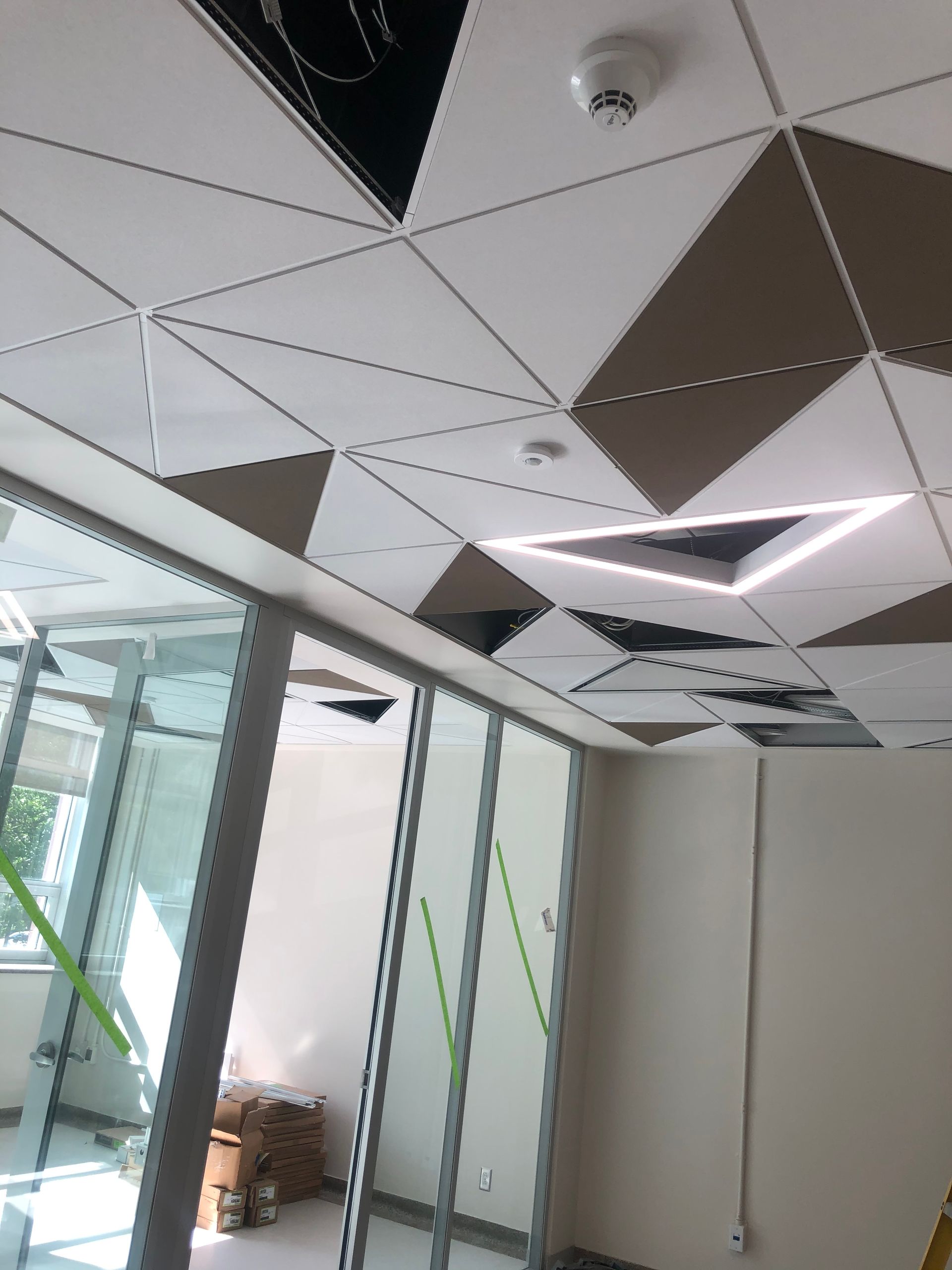 Plafond de bureau avec carreaux géométriques blancs, marron et noirs, éclairage encastré et cloison en verre.