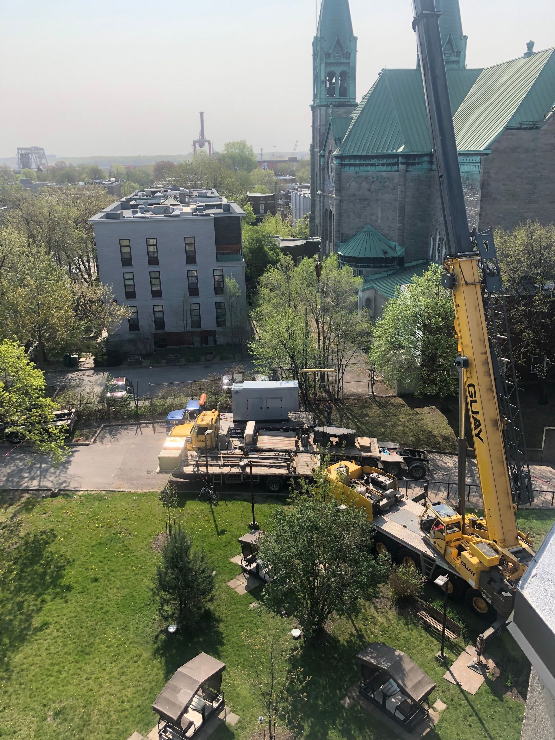 Une grande grue jaune près d'une église en pierre et de bâtiments gris. Matériaux de construction au sol, herbe verte.