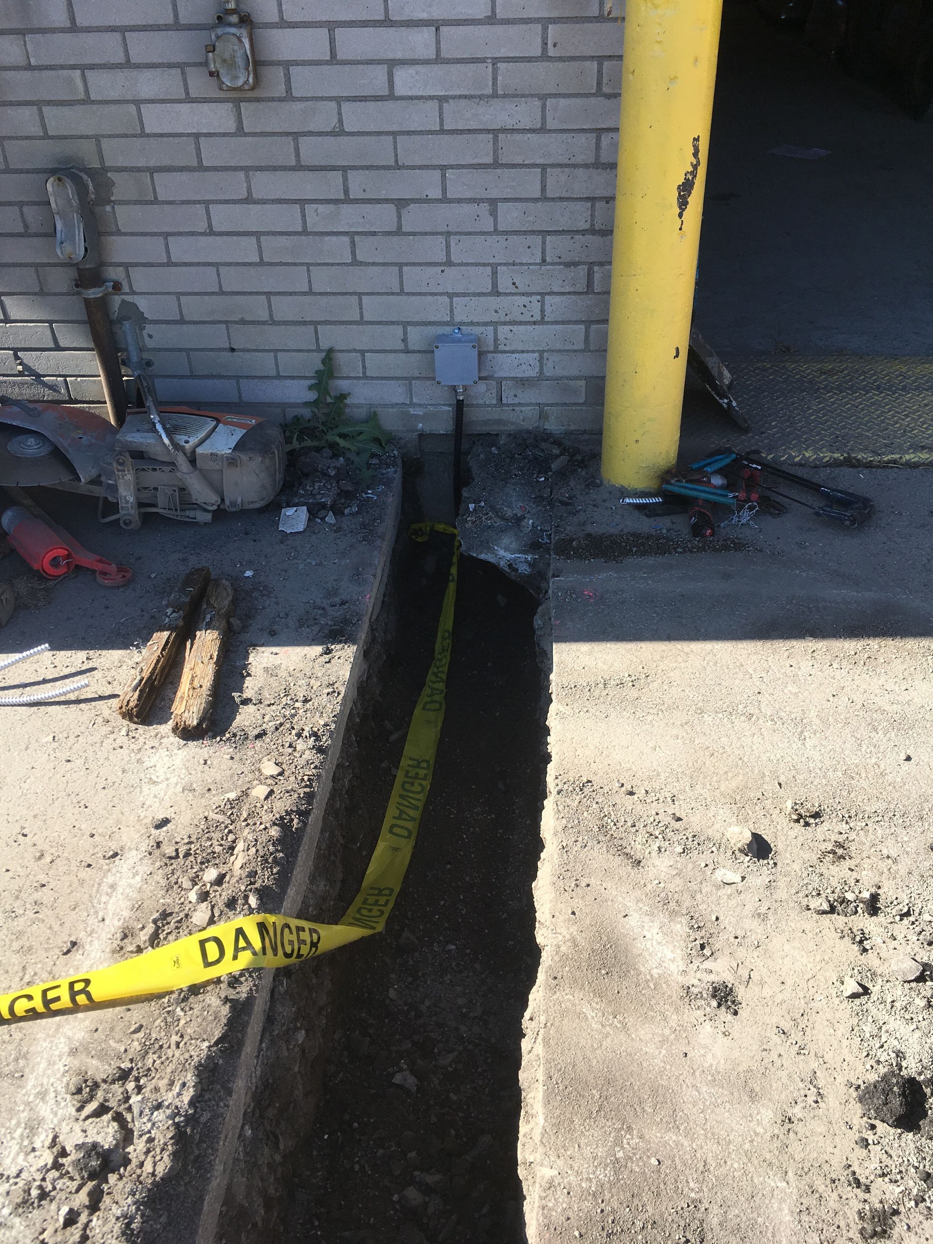 Tranchée creusée dans le béton avec ruban d'avertissement jaune ; à côté d'un mur de briques et d'un poteau jaune ; pour travaux de services publics.