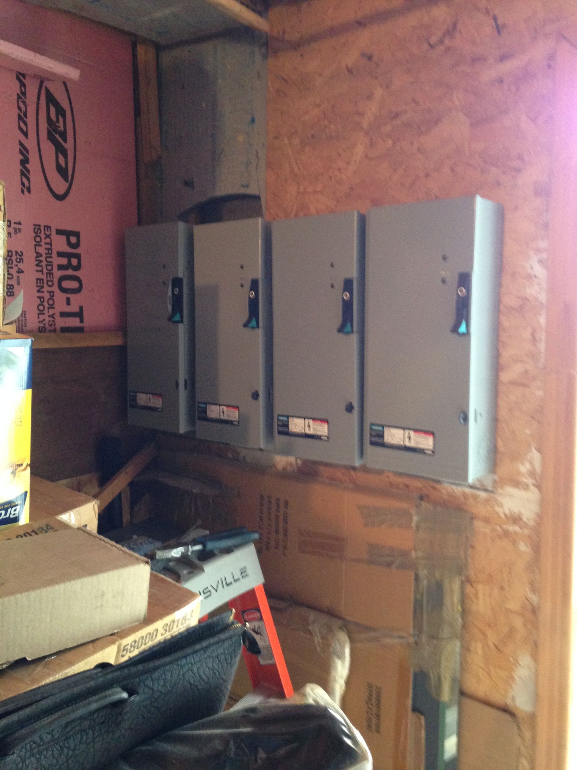 Quatre panneaux électriques gris montés sur un mur en bois dans une zone de stockage encombrée.