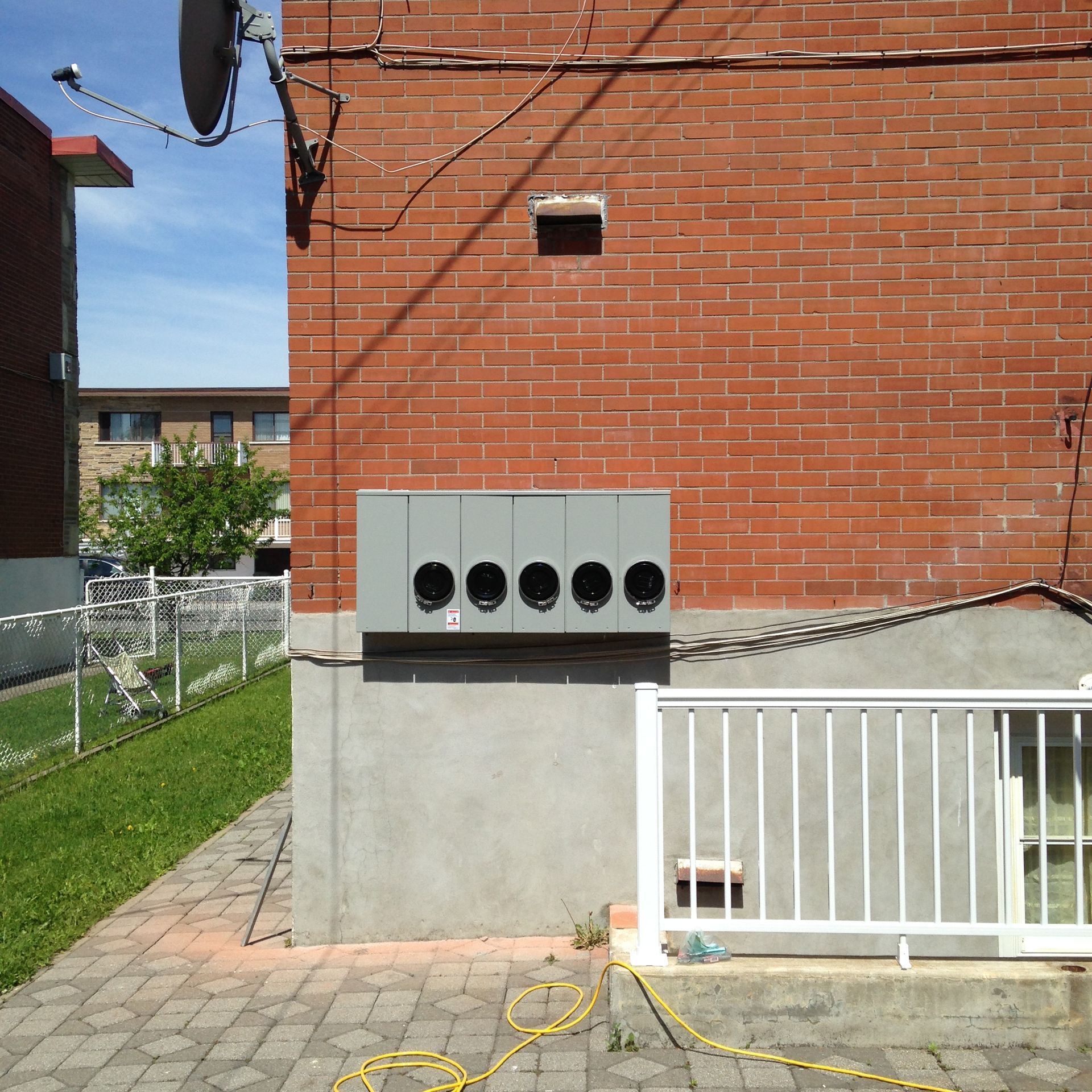 Un boîtier multimètre gris sur un bâtiment en briques rouges avec une clôture blanche et une zone pavée.