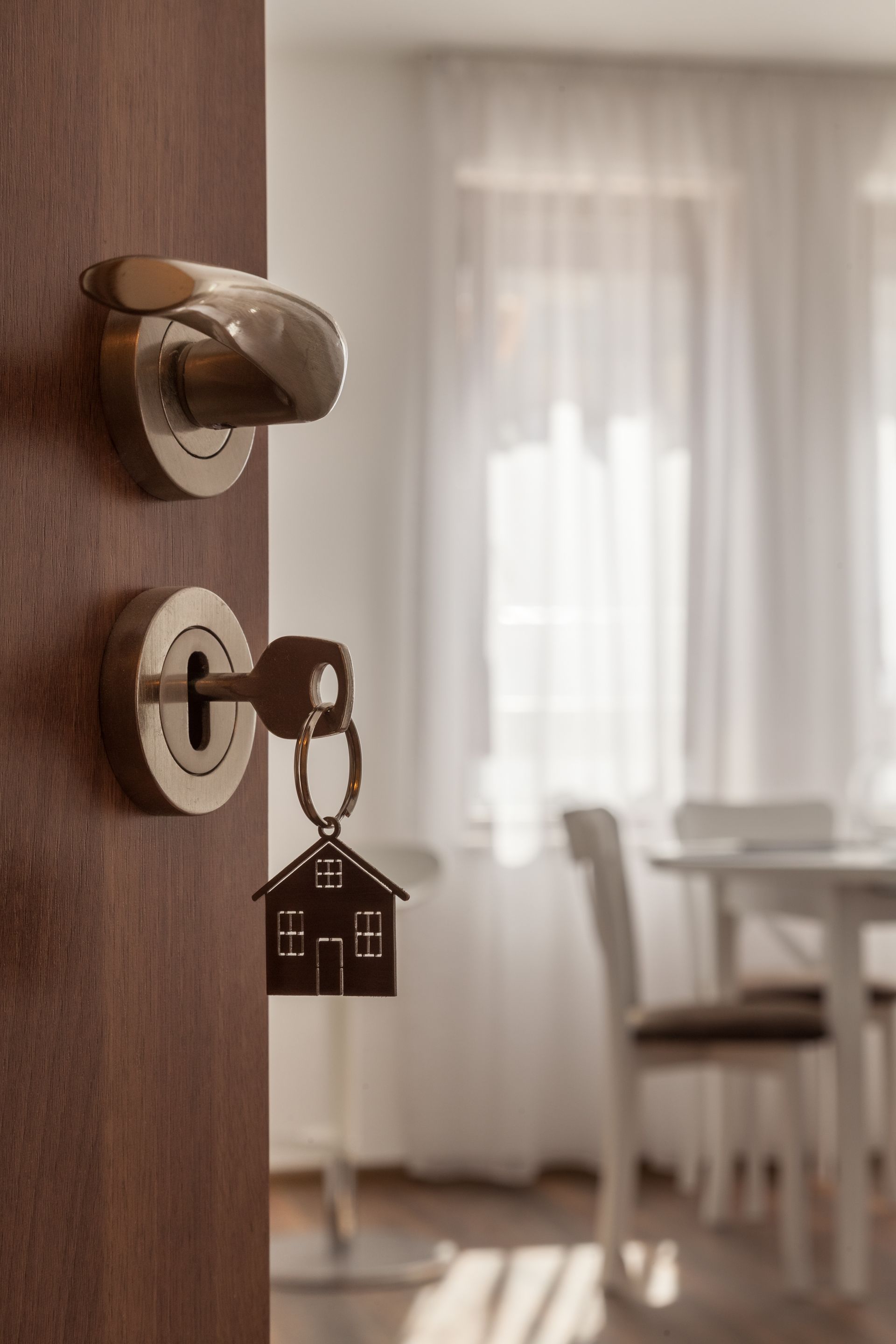 Door ajar, key in lock with house-shaped keychain, revealing a dining area with table and chairs.