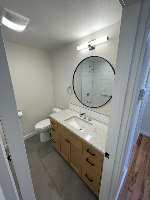 Small bathroom with white walls, vanity sink, round mirror, toilet, and gray tile floor.