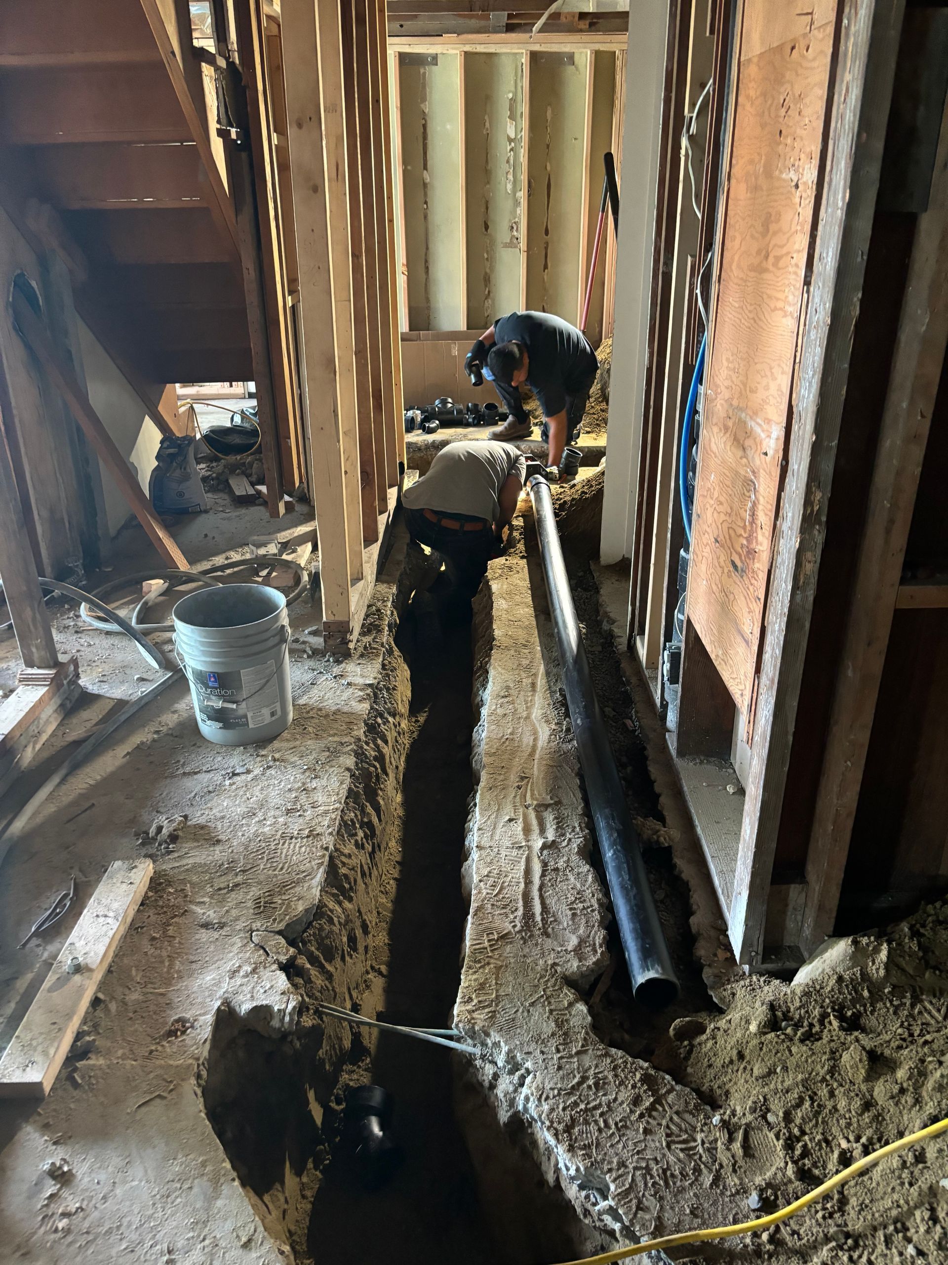 Worker installing underground piping in a wooden-framed construction site with a trench and buckets
