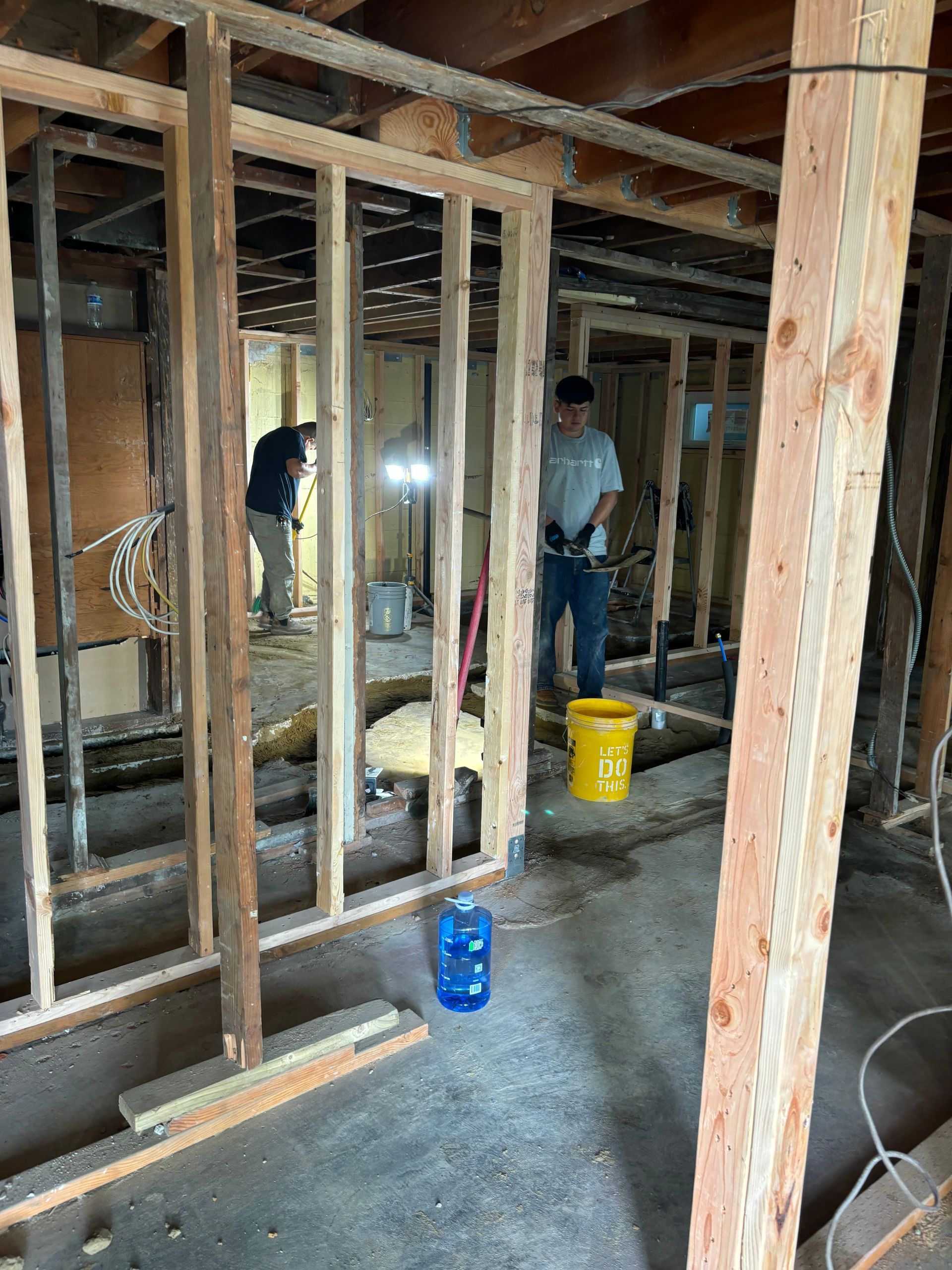 Unfinished basement framed with exposed studs; two workers stand among tools and buckets on the concrete floor.