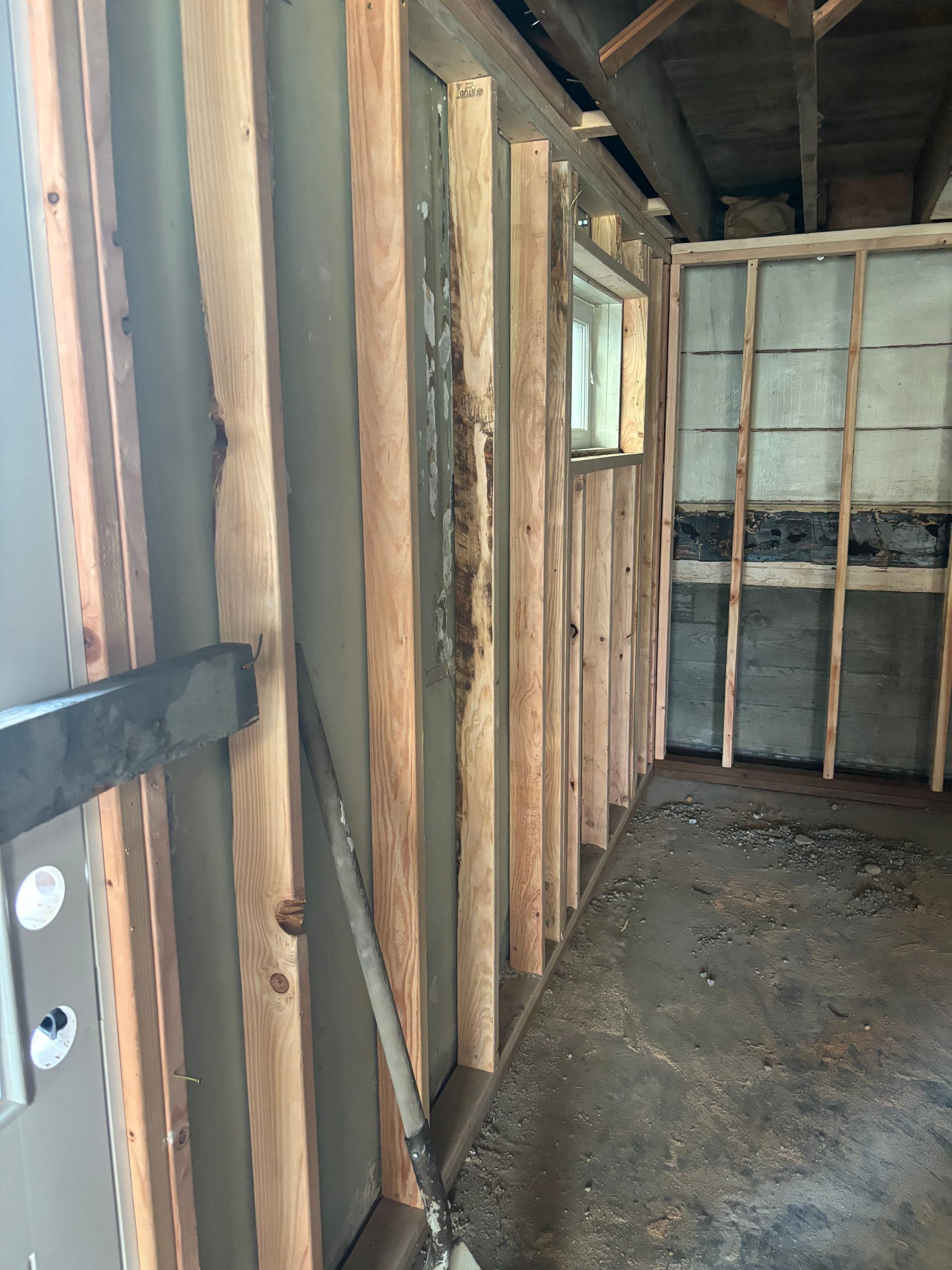 Interior of an unfinished wooden shed with exposed studs, a small window, and a dirt floor.