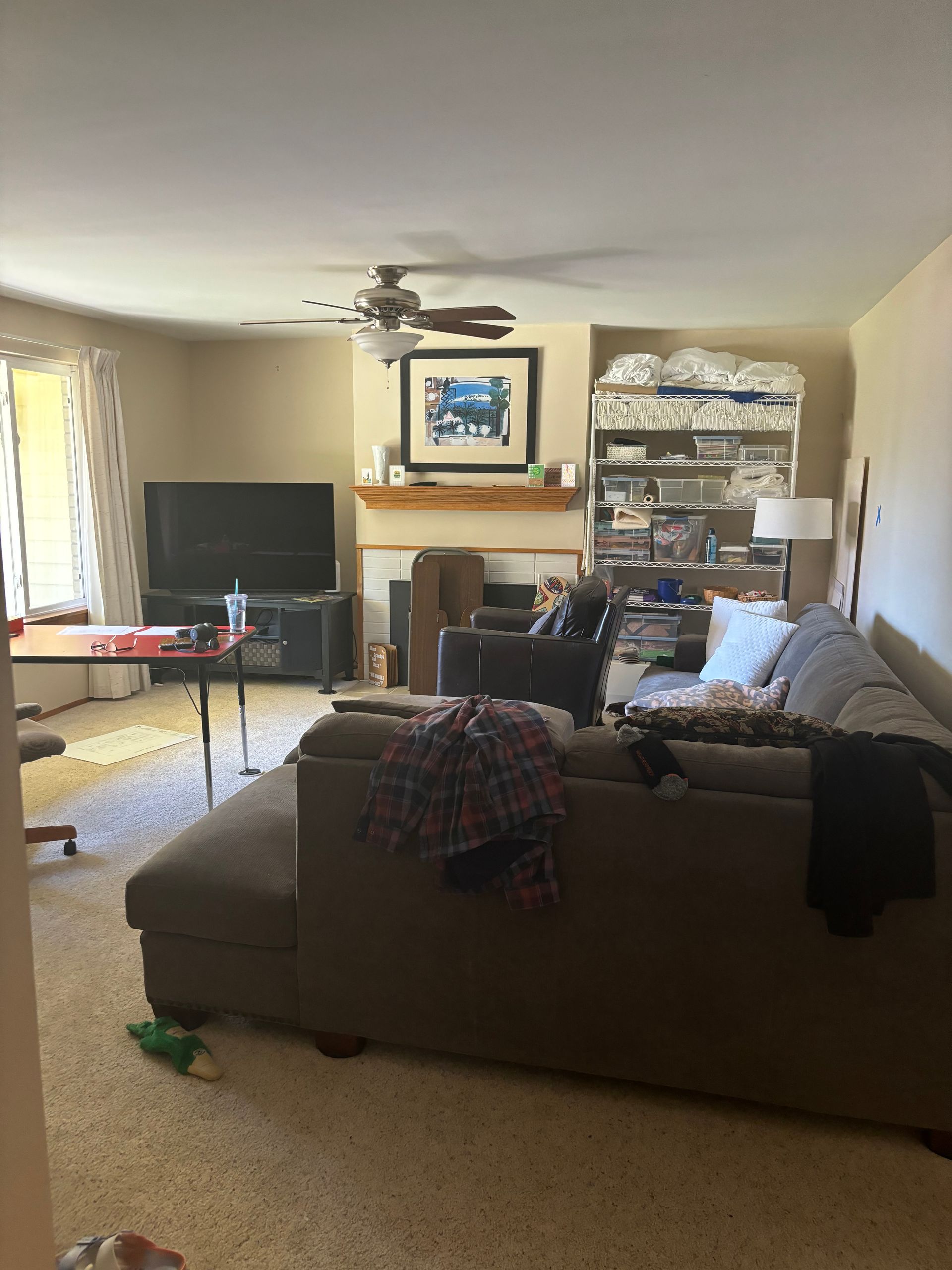 Living room with sectional sofa, TV, fireplace, shelves, and a ceiling fan on beige carpet.