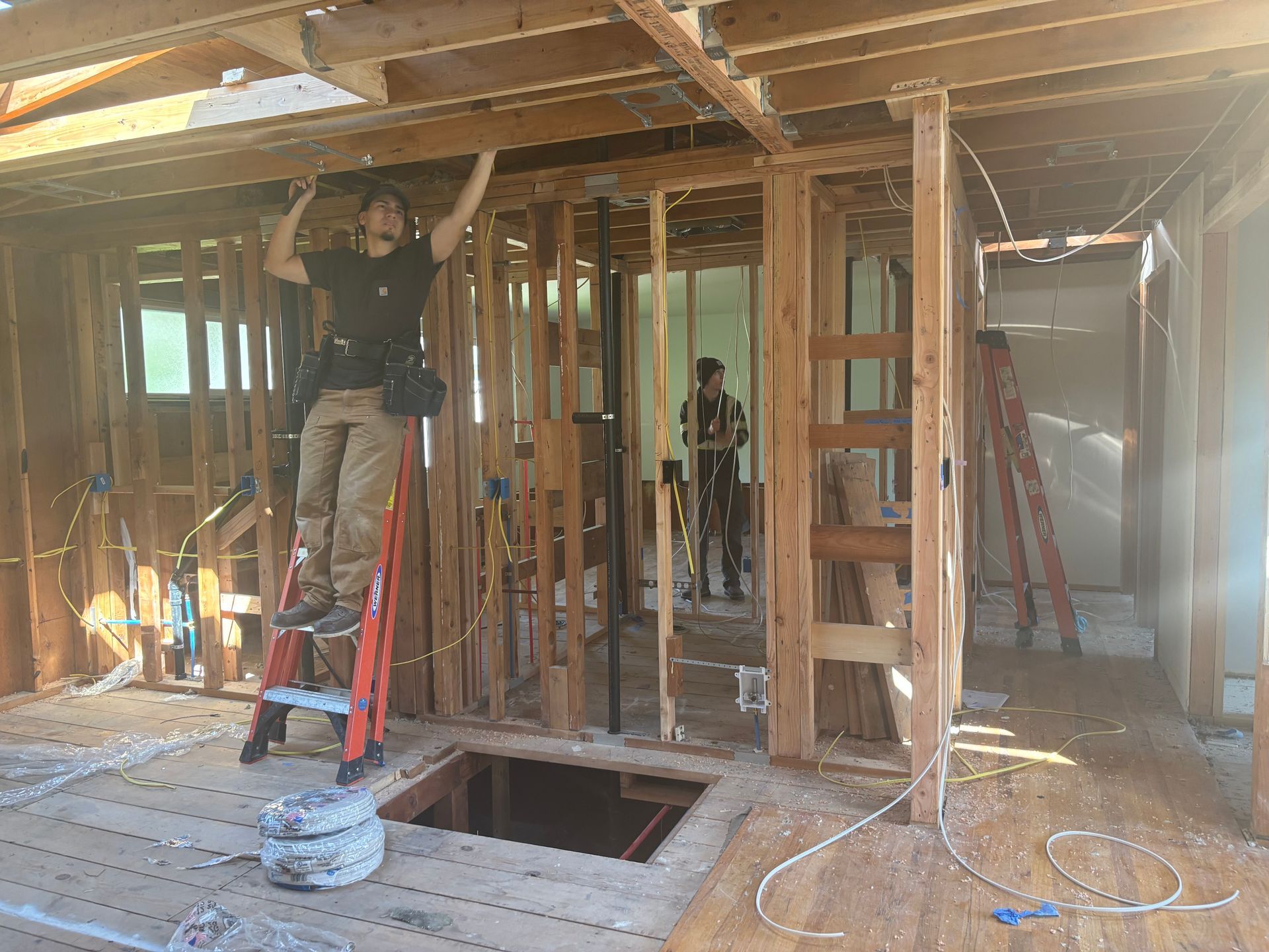Two workers framing a house interior under construction, with exposed studs and a ladder.