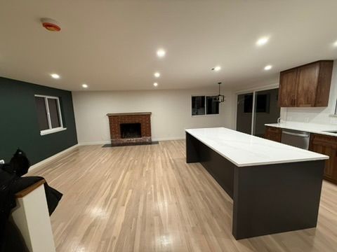 Empty modern kitchen and living area with wood floors, island, recessed lights, and a brick fireplace.