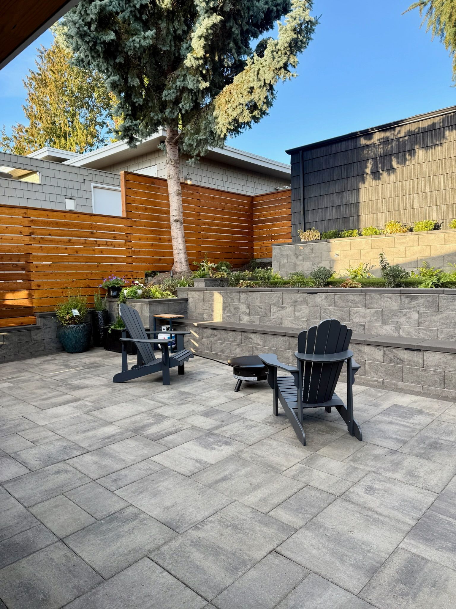 Patio with stone pavers, black chairs, raised planter beds, wooden fence, and tall trees at sunset