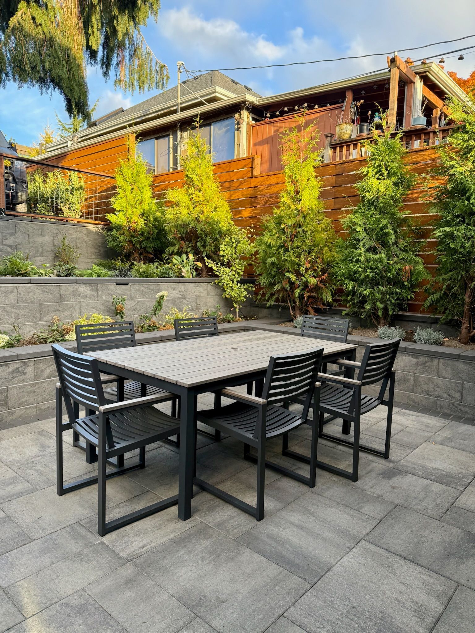 Outdoor patio with black table and chairs, stone paving, and a wooden house behind lush green shrubs