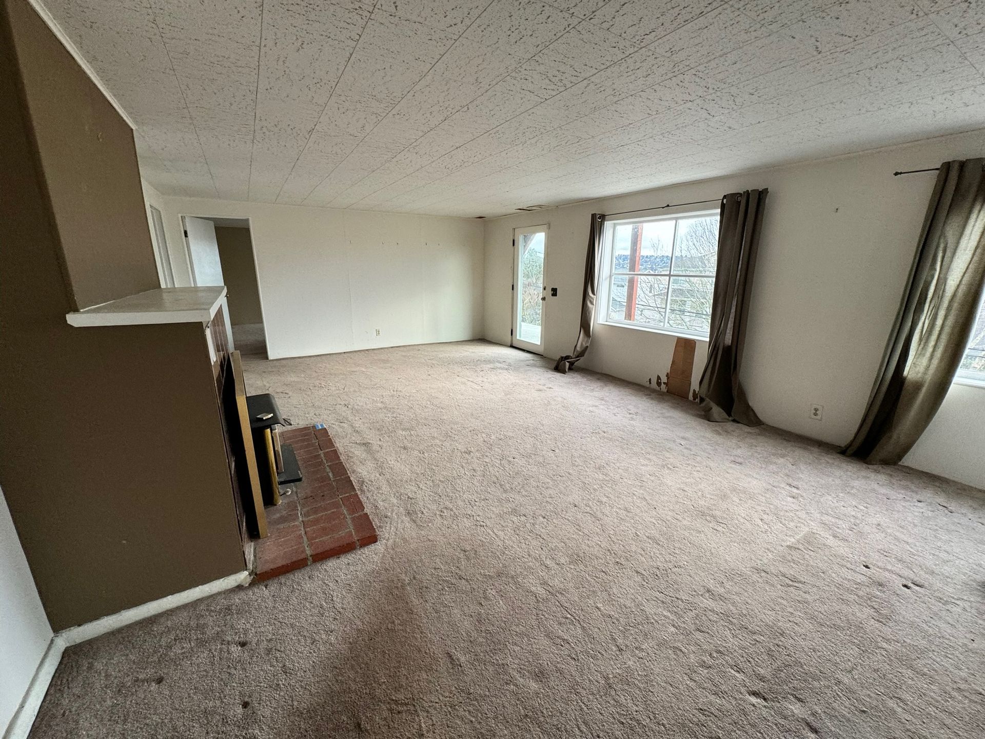 Empty beige carpeted living room with fireplace and large windows with curtains.