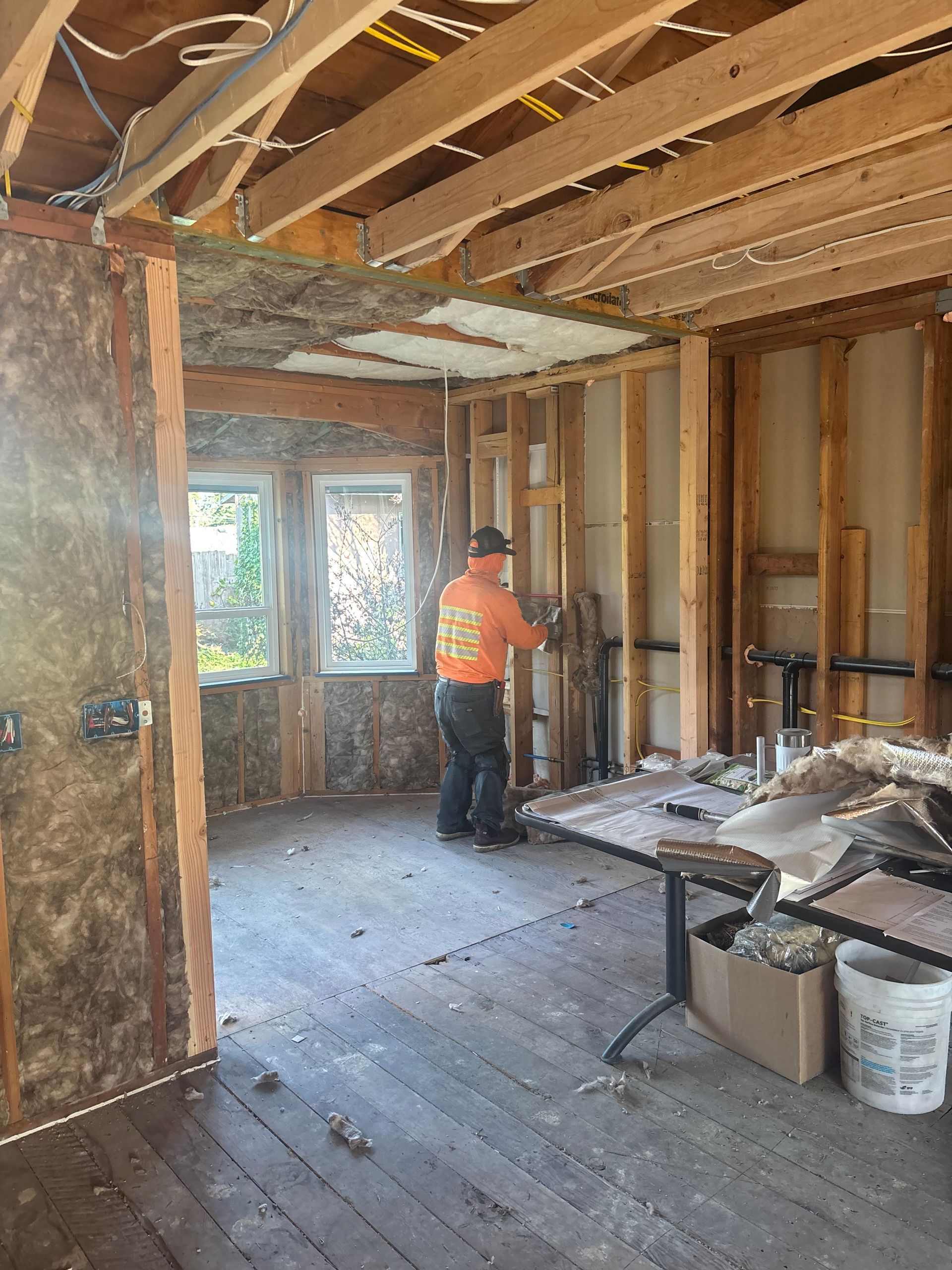 Partially renovated room with exposed studs and beams; worker in orange shirt stands near a table of tools and materials.