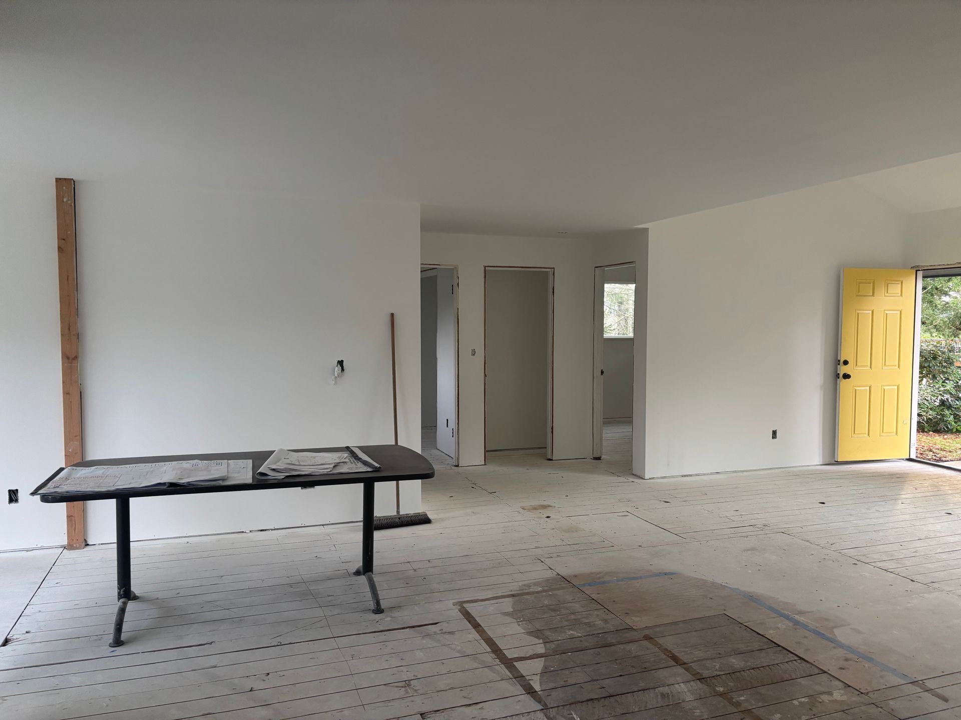 Empty unfinished room with white walls, tiled floor, and a work table near a yellow front door.