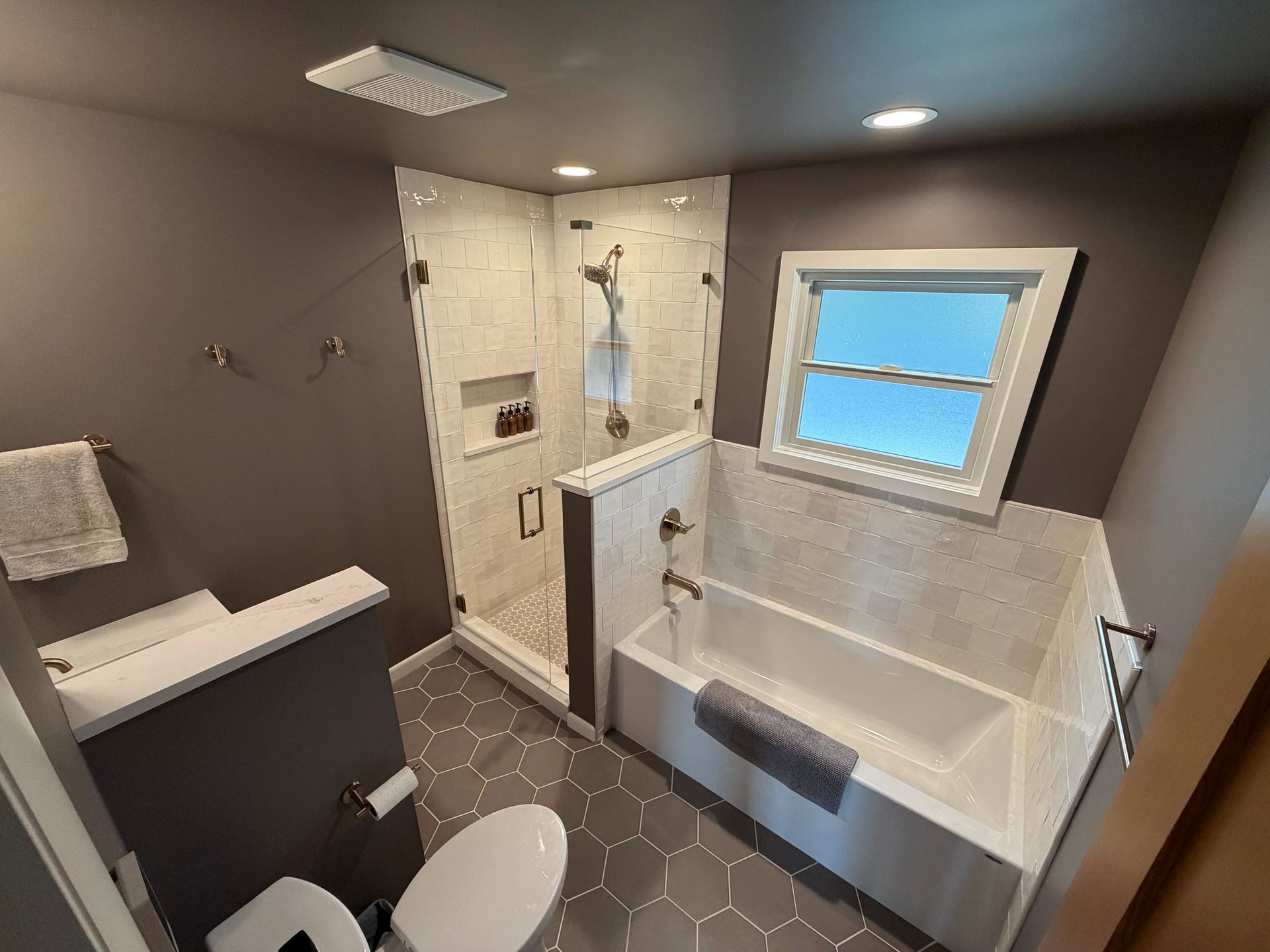 Modern gray bathroom with walk-in shower, white tub, and small frosted window