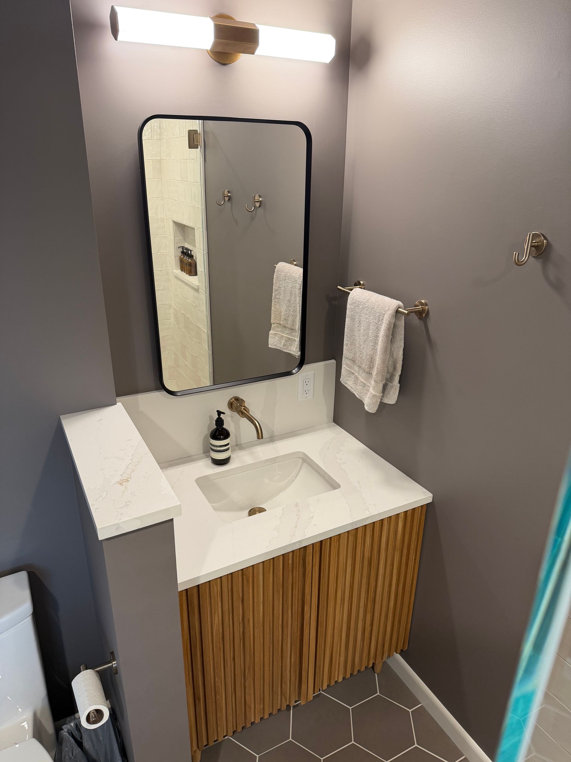 Small gray bathroom with vanity sink, mirror, towel ring, and wood cabinet under bright light.
