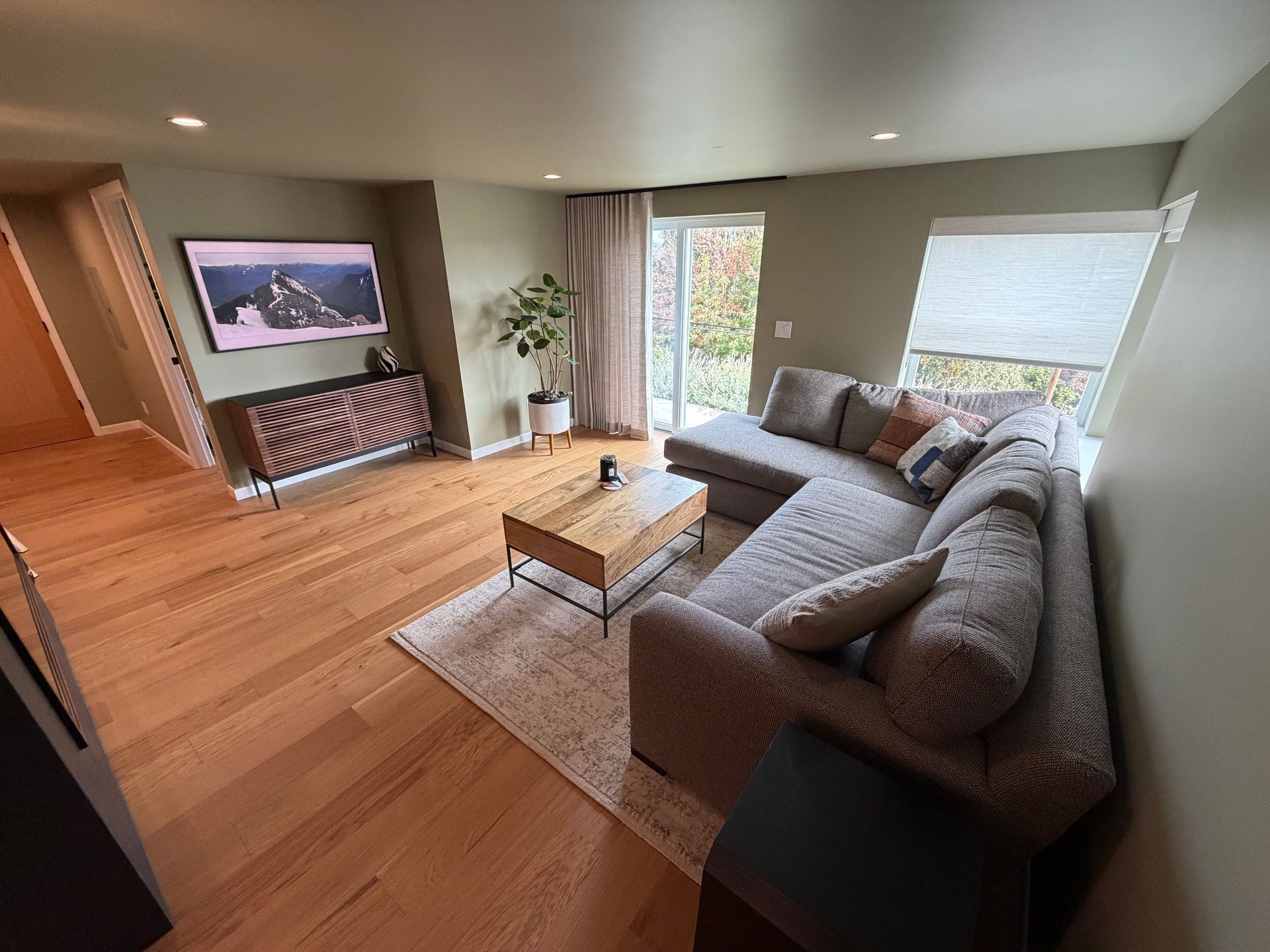 Bright living room with gray sectional, wooden coffee table, TV, and large windows with city view.