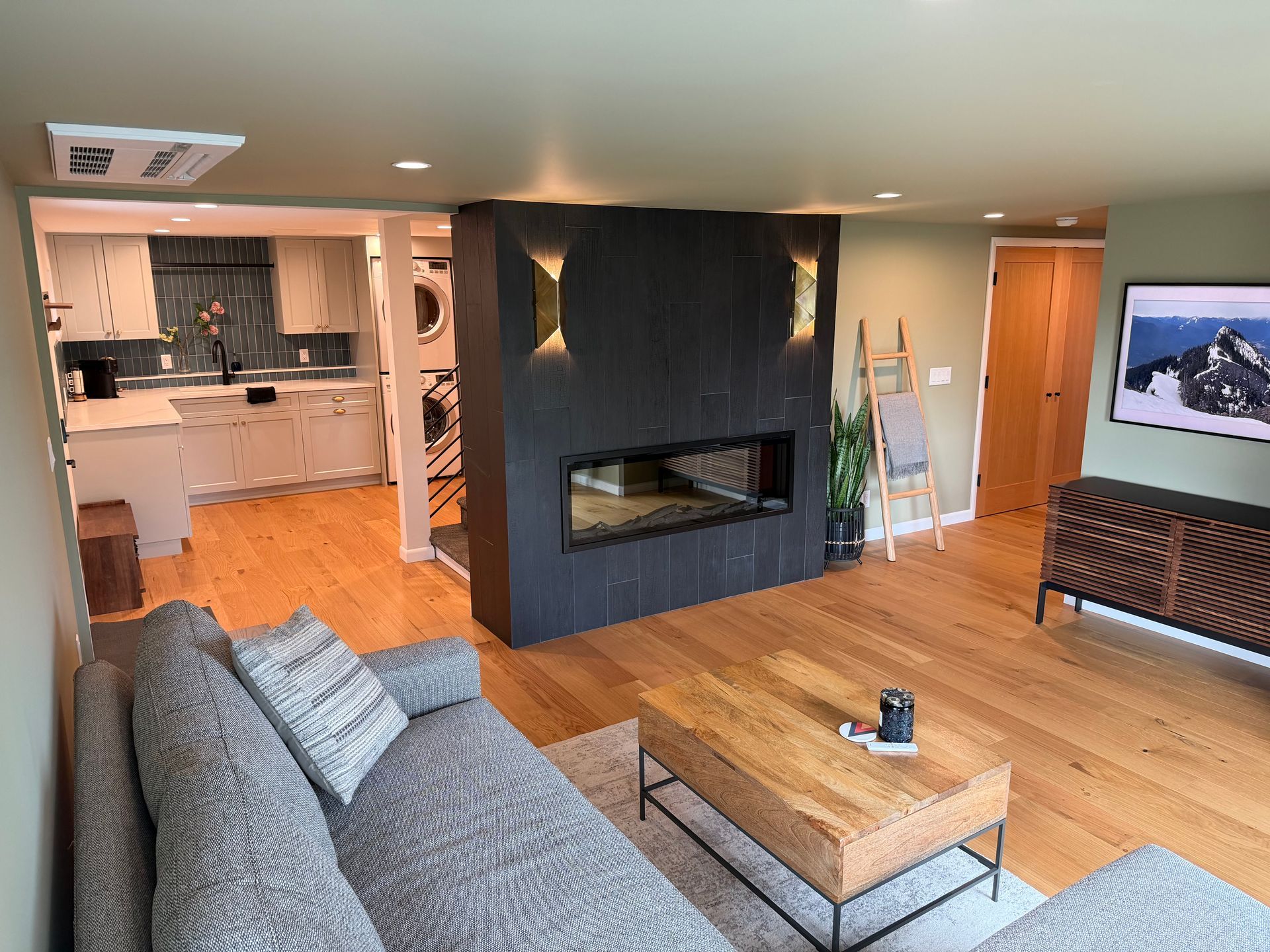 Modern living room with wood floors, gray sectional, black fireplace, and open kitchen in the background