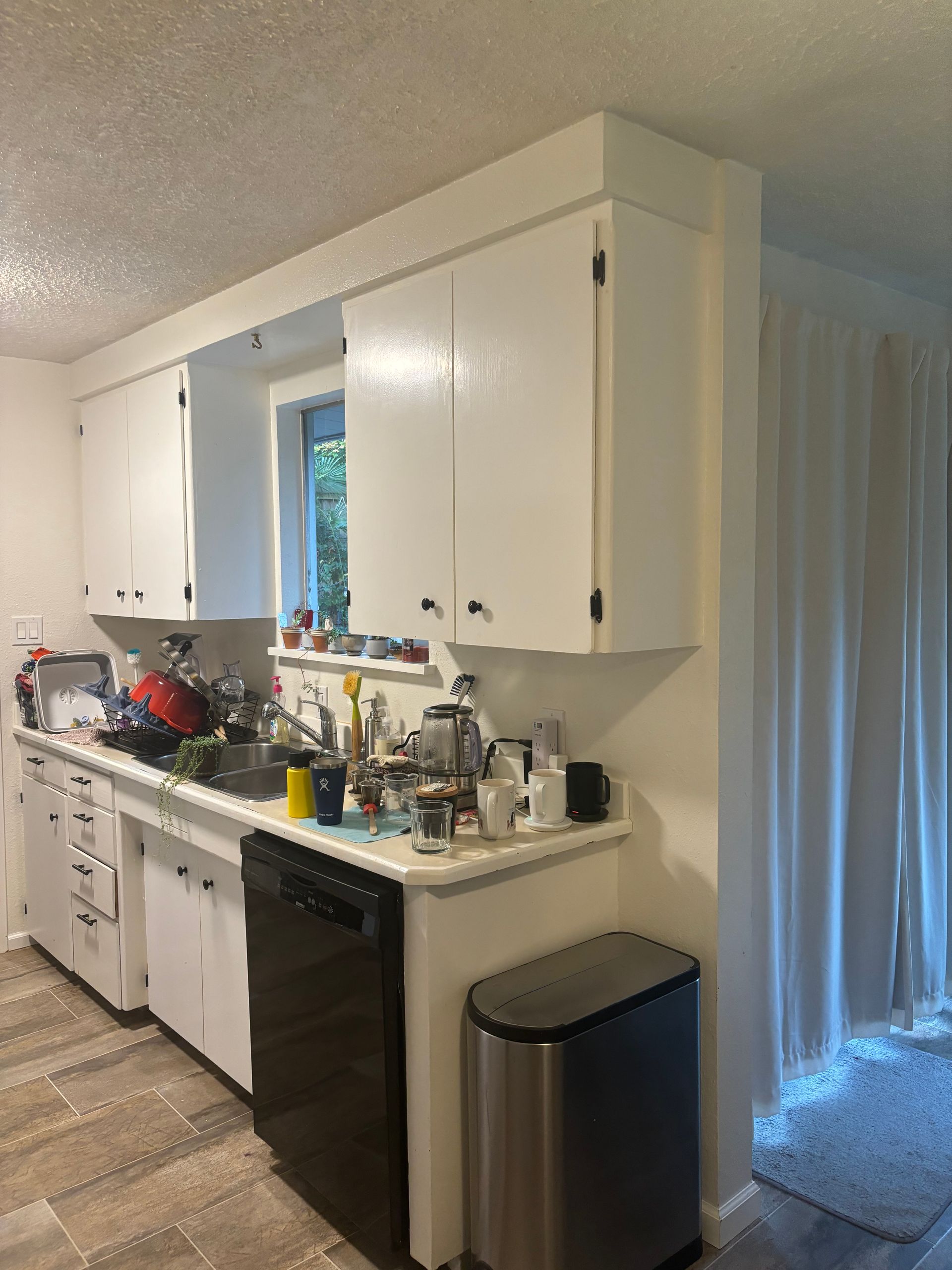 Kitchen with white cabinets, cluttered countertop, black dishwasher, and stainless trash can beside a doorway