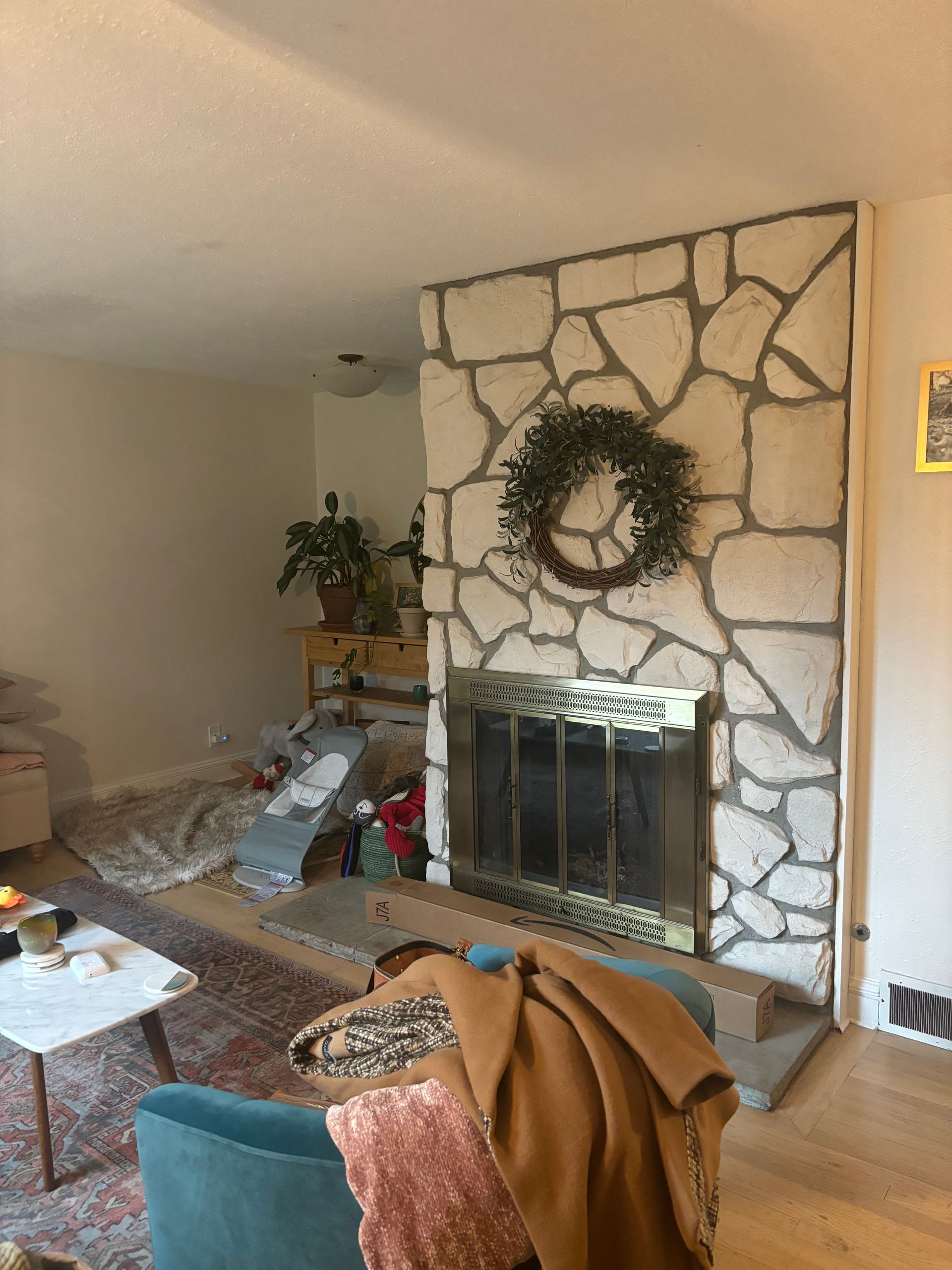 Living room with stone fireplace, wreath, teal chair, and a couch by a window