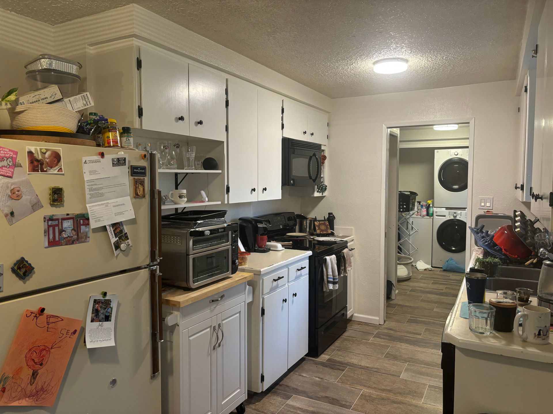 Narrow kitchen with white cabinets, fridge, and appliances, viewed toward a laundry area in the back.