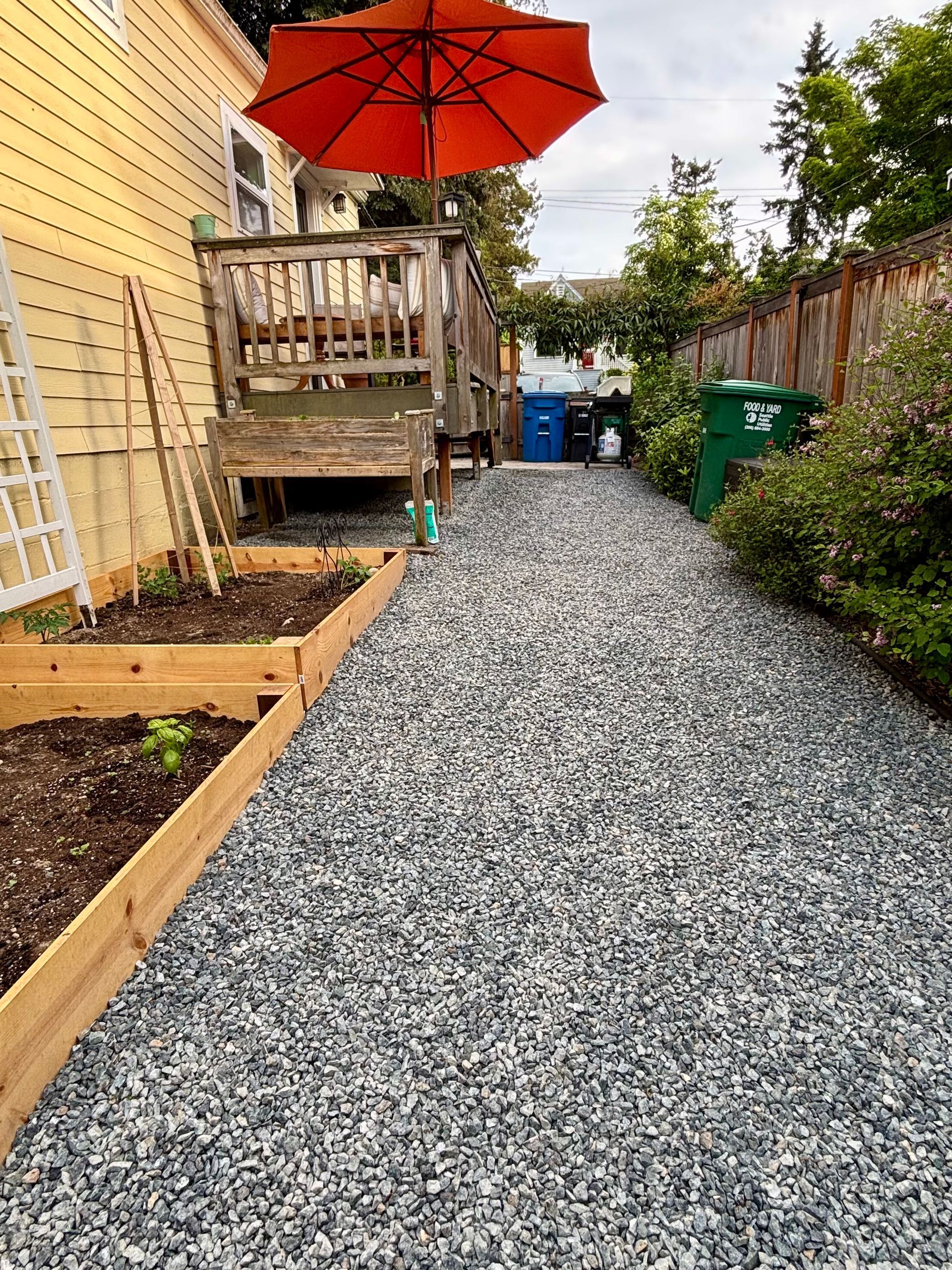 Gravel backyard path beside a wooden deck with an orange umbrella and raised garden beds, bordered by fences and shrubs