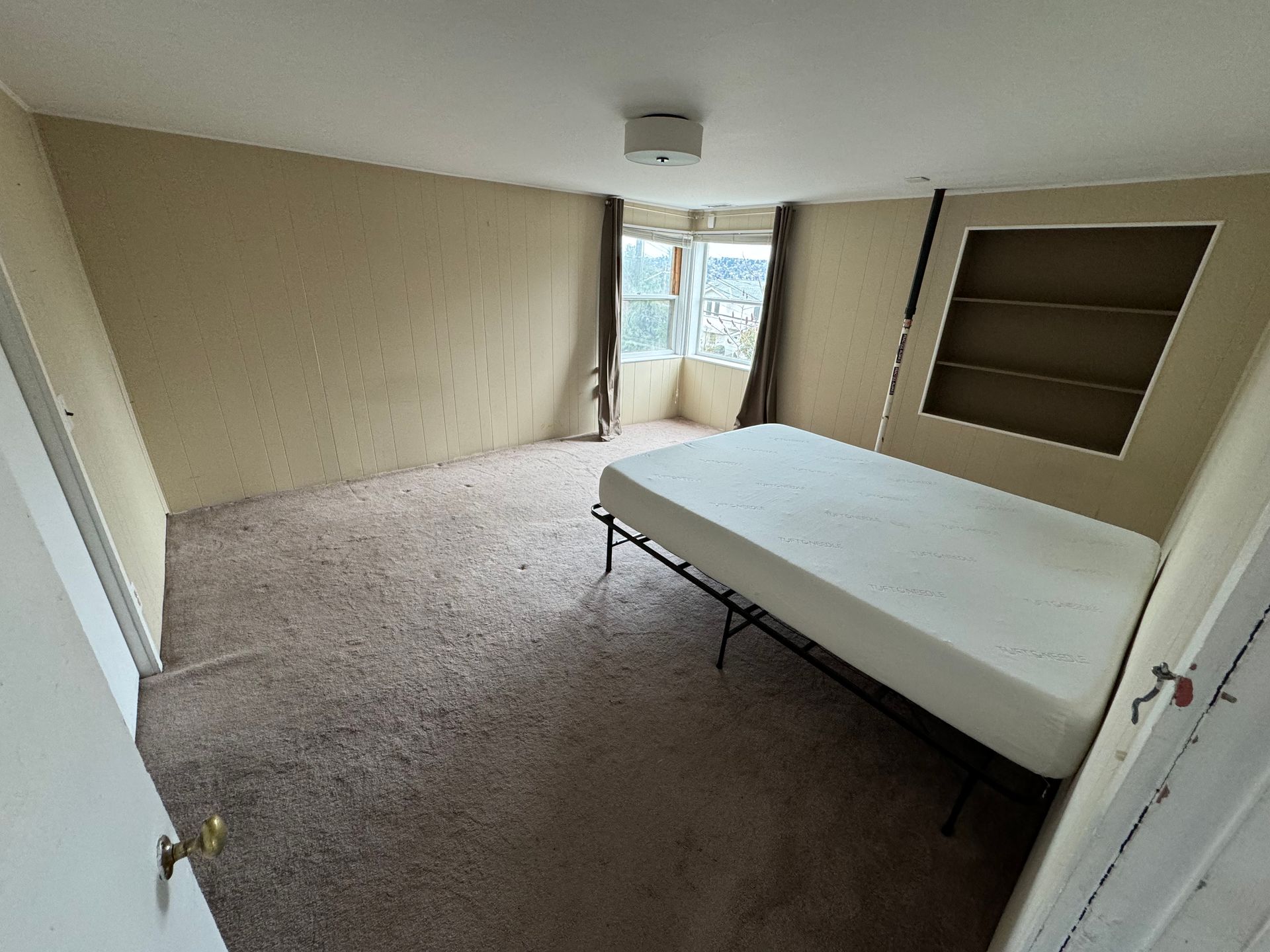 Empty beige bedroom with carpet, a bed frame, built-in shelves, and a window at the far end.