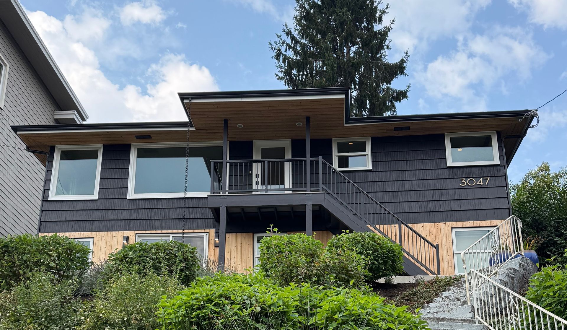 Two-story gray house with wood accents, front porch, and landscaped shrubs under a cloudy sky