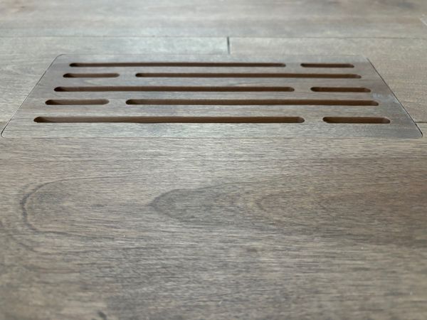 Grille d'aération de plancher en bois avec fentes horizontales, encastrée dans un plancher à grain de bois.