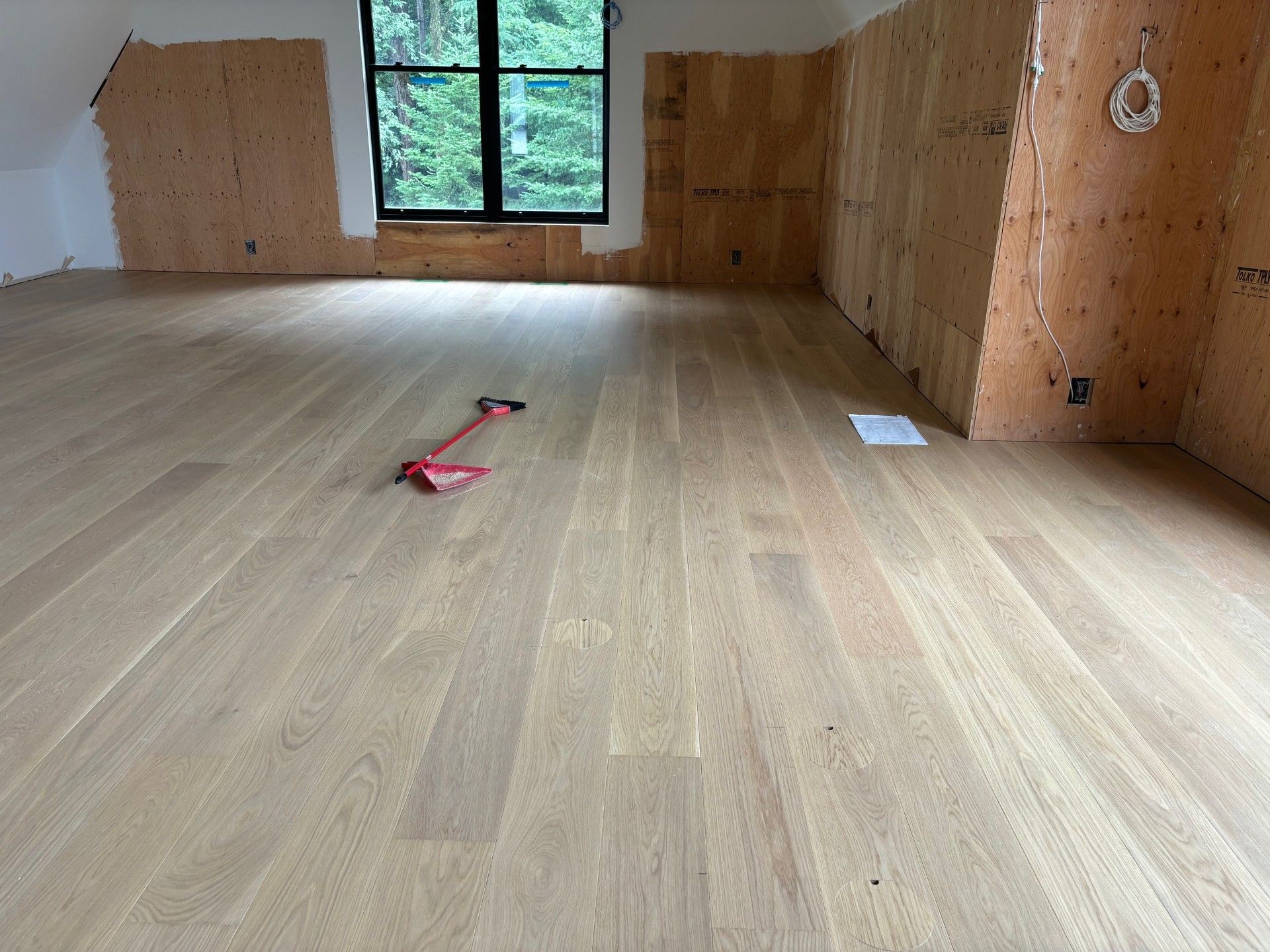 Parquet clair récemment installé dans une pièce avec une fenêtre, des murs non finis et un pied de biche rouge.